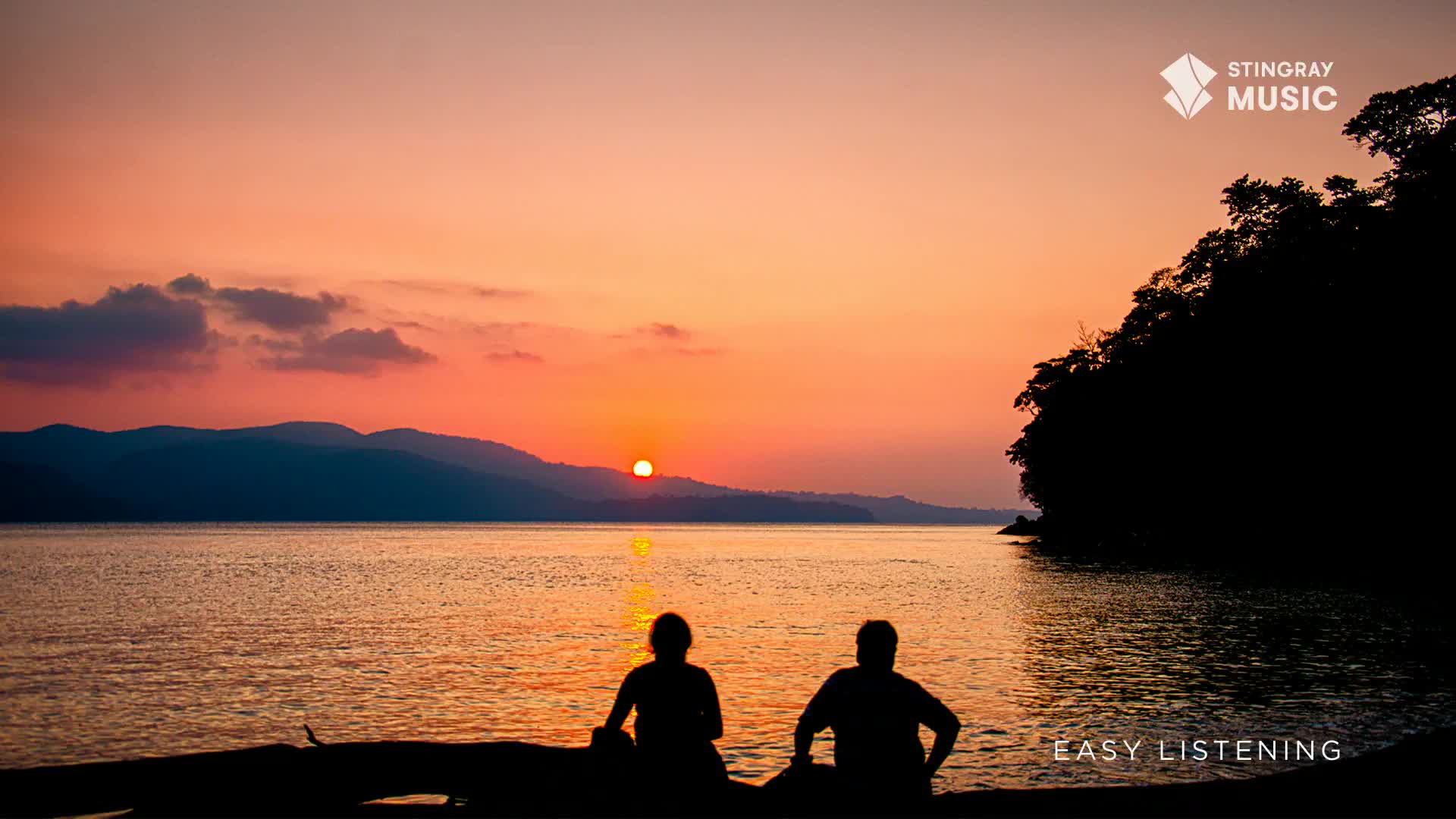 The setting sun casts a warm glow over the water, reflecting off the surface. Two figures sit on a log, silhouetted against the vibrant sky, enjoying the easy listening vibe.
The setting sun casts a warm glow over the water, reflecting off the surface. Two figures sit on a log, silhouetted against the vibrant sky, enjoying the easy listening vibe.