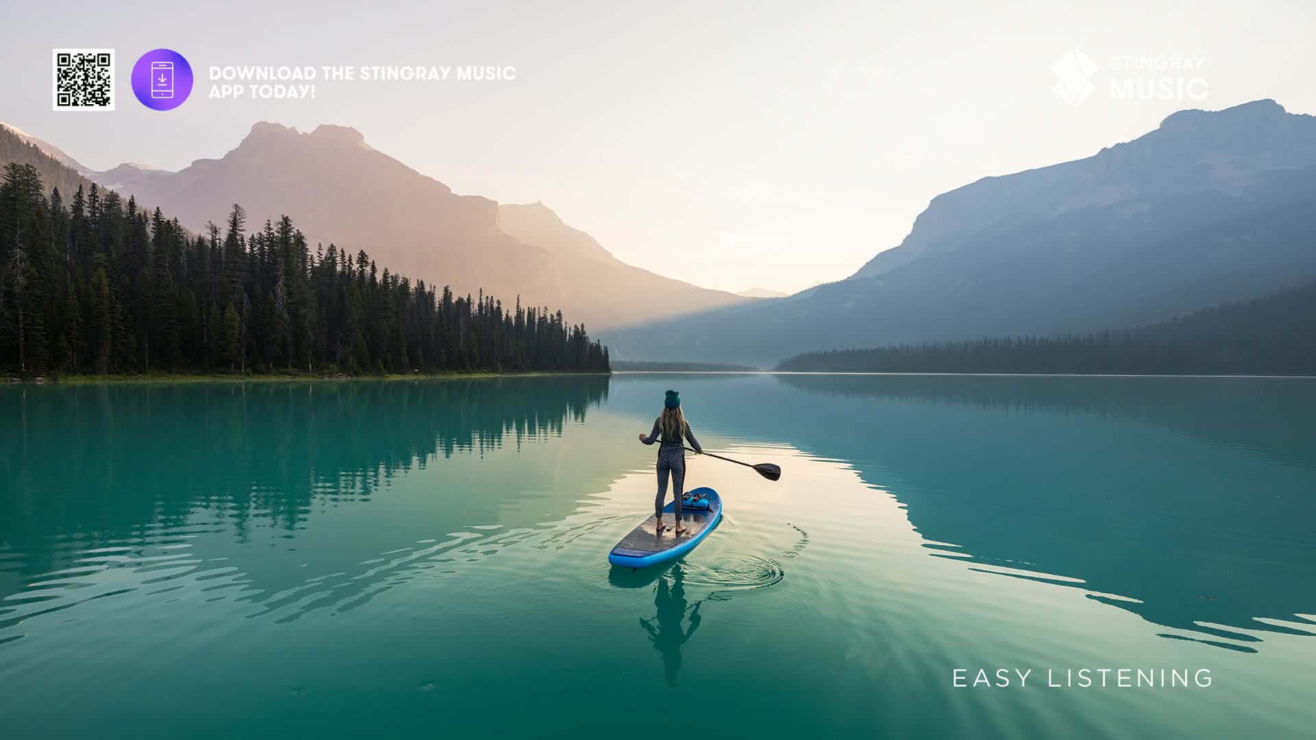 A person stands on a paddleboard, gracefully gliding across the turquoise water of a Canadian lake. The mountains rise in the distance, and the scene feels peaceful, like the kind of easy listening music you'd play while enjoying the view.
