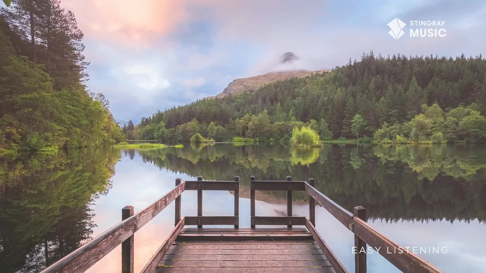 The lake's surface mirrors the trees and the sky, a perfect reflection. A wooden dock juts out, inviting a moment of quiet contemplation, the kind of scene that might accompany Stingray Easy Listening from Canada.

