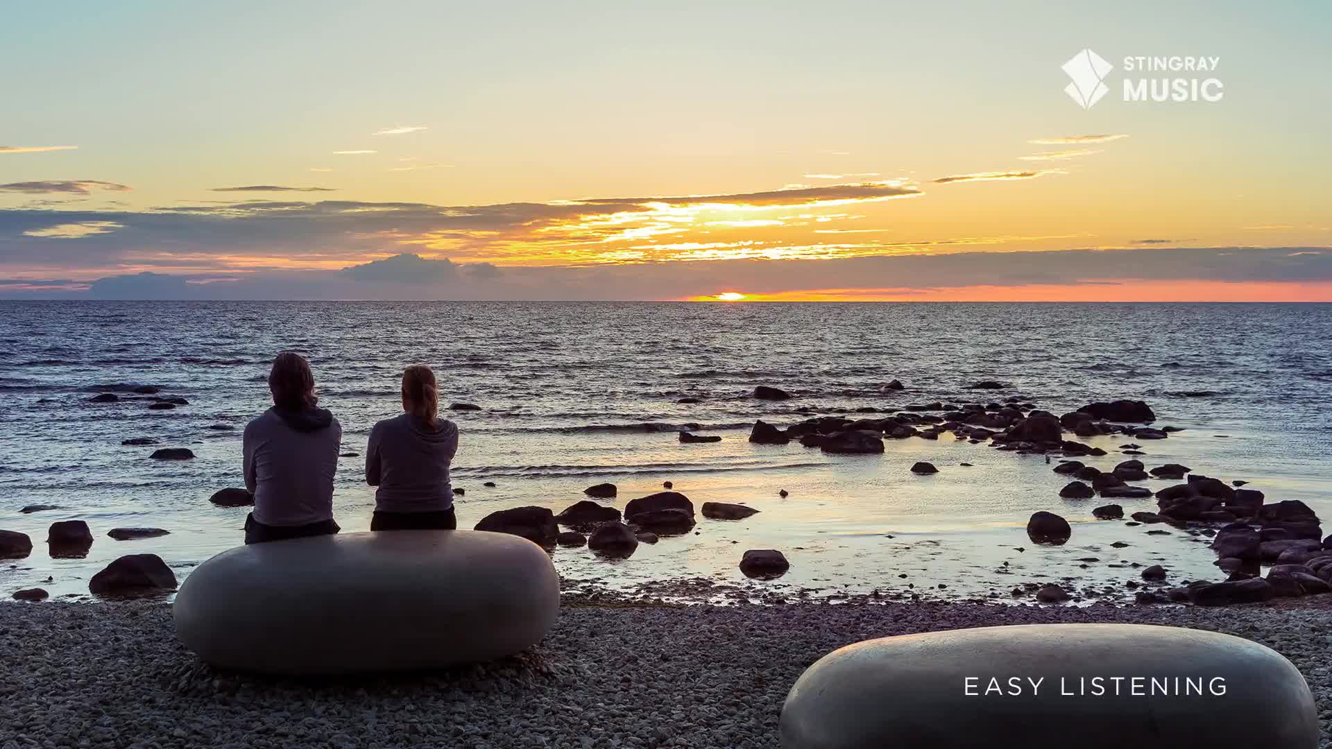 Two figures sit on a smooth, grey bench, facing the vast ocean as the sun dips below the horizon. The sky above is painted with soft hues of orange and pink, reflecting on the gentle waves.