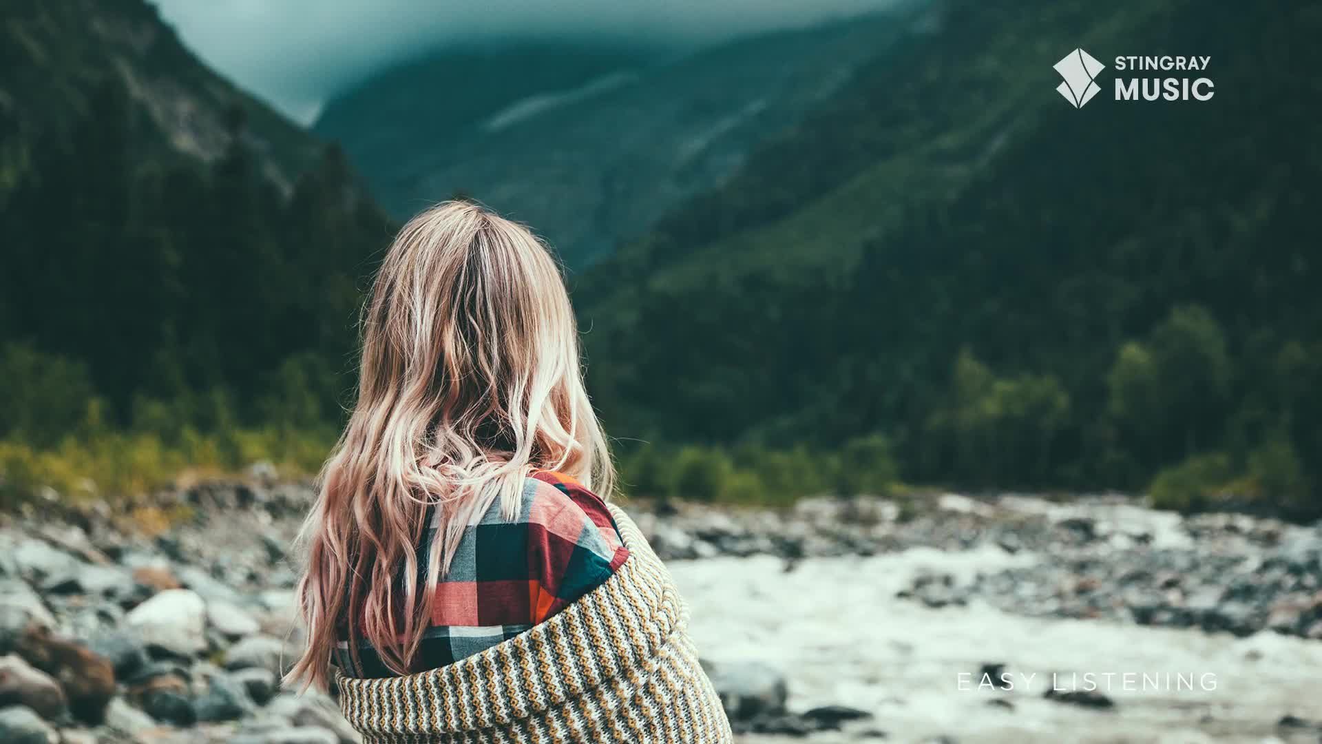 A young woman with long, blonde hair stands by a rocky riverbank, her back to me. The water rushes past, a cool grey against the lush green of the Canadian mountains.