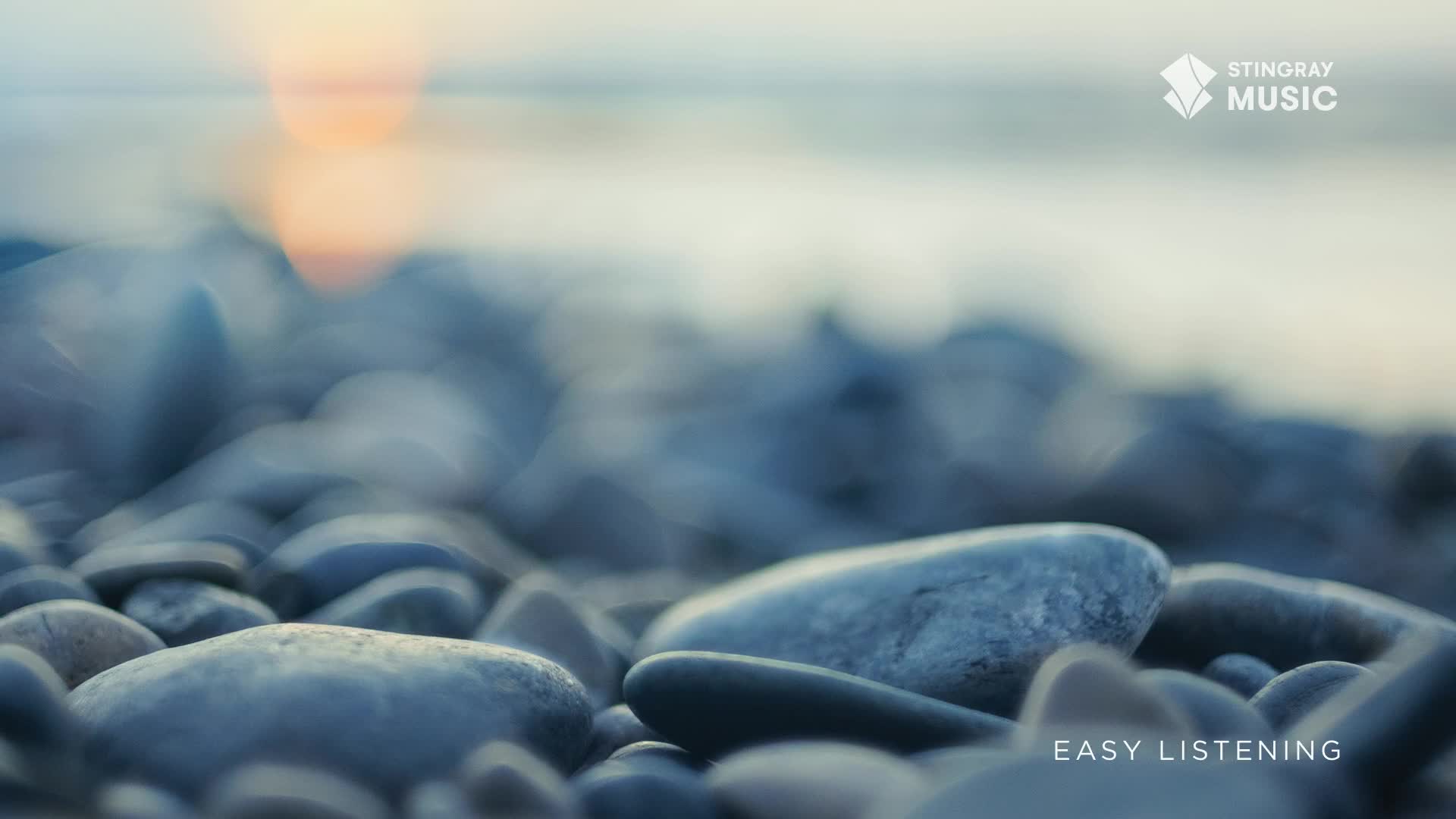 Smooth, grey stones fill the foreground, leading to a hazy horizon where the sun dips low. The gentle lapping of water on a Canadian shore seems to accompany the soft glow.