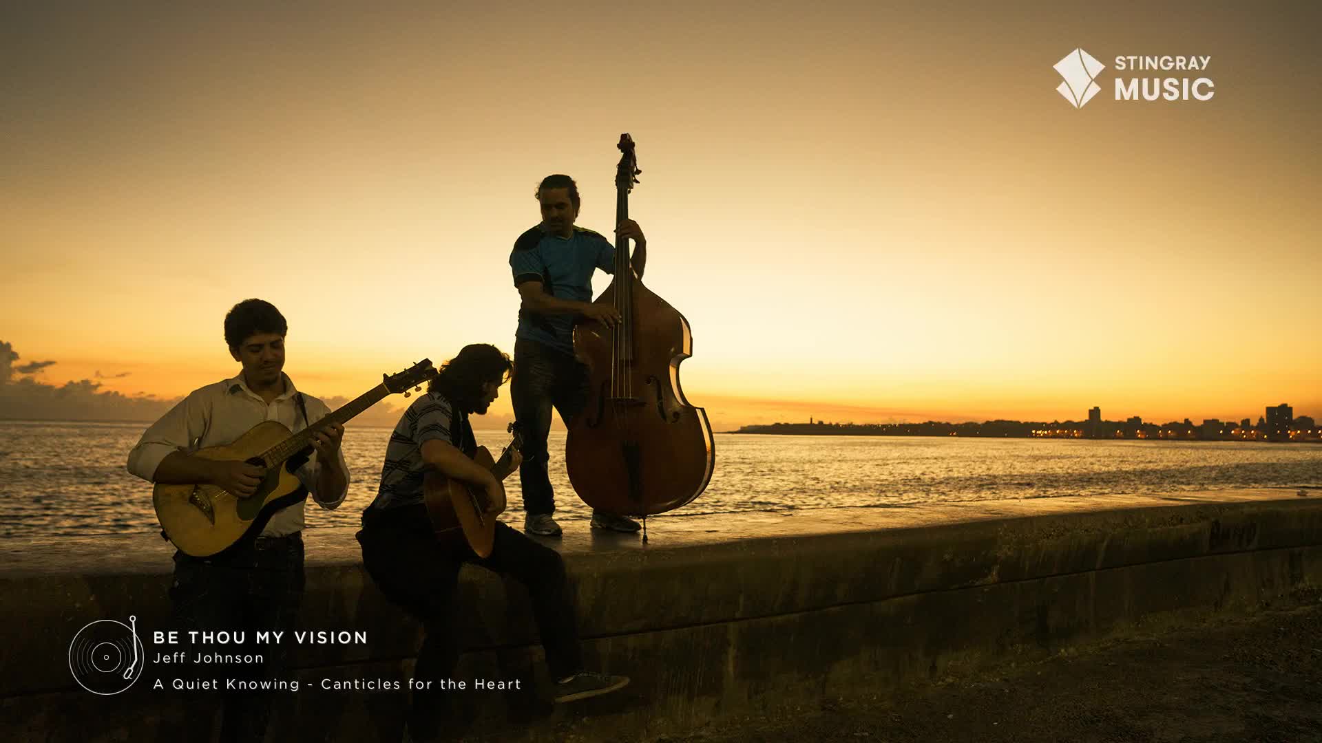 Three musicians play by the water as the sun sets over a distant Canadian city skyline. A guitarist strums his instrument while a second guitarist sits playing, and a third man stands behind a double bass.