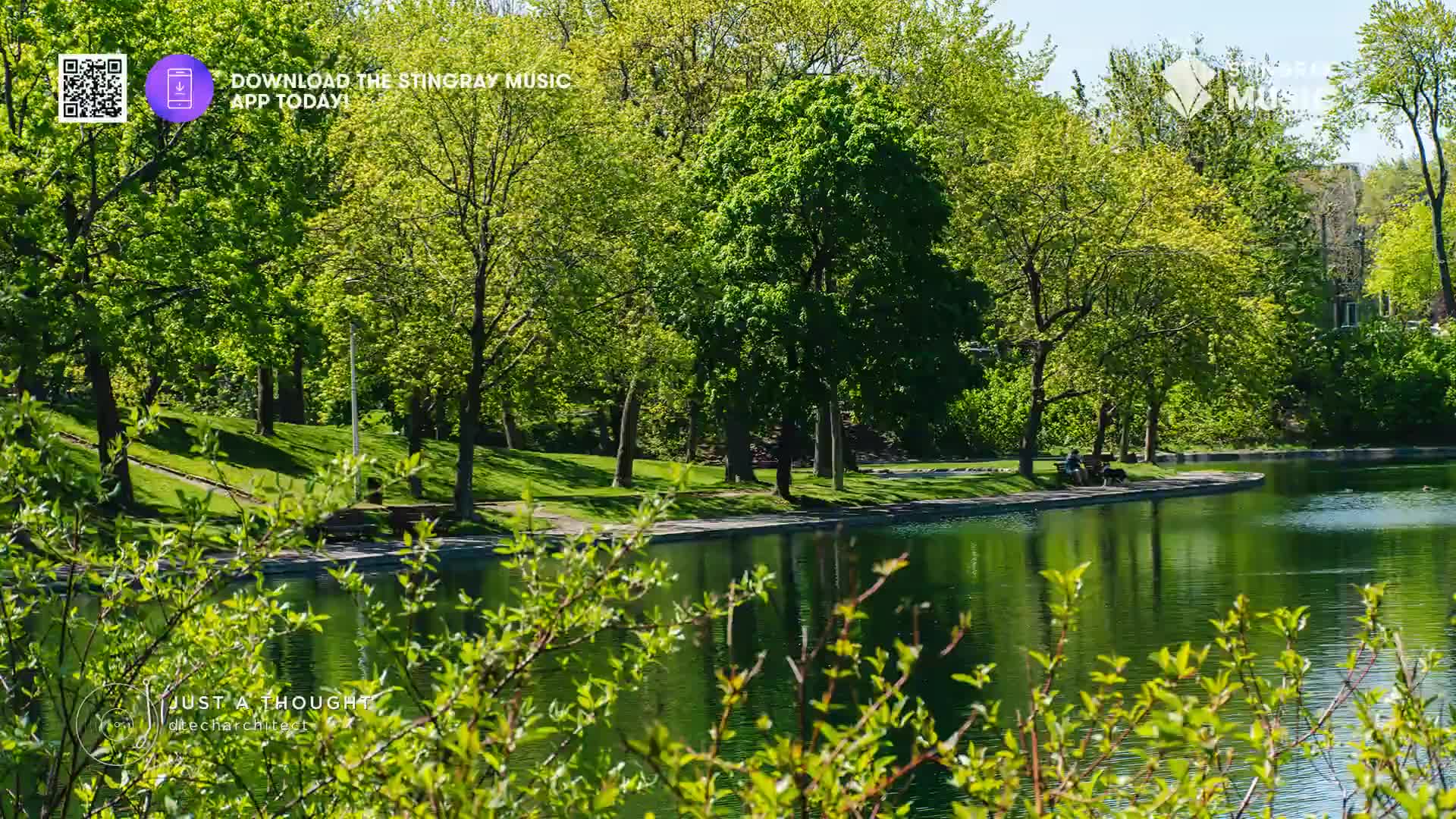 A couple sits on a park bench by the water's edge, their dog resting at their feet. The lush green trees and calm lake create a peaceful Canadian scene.