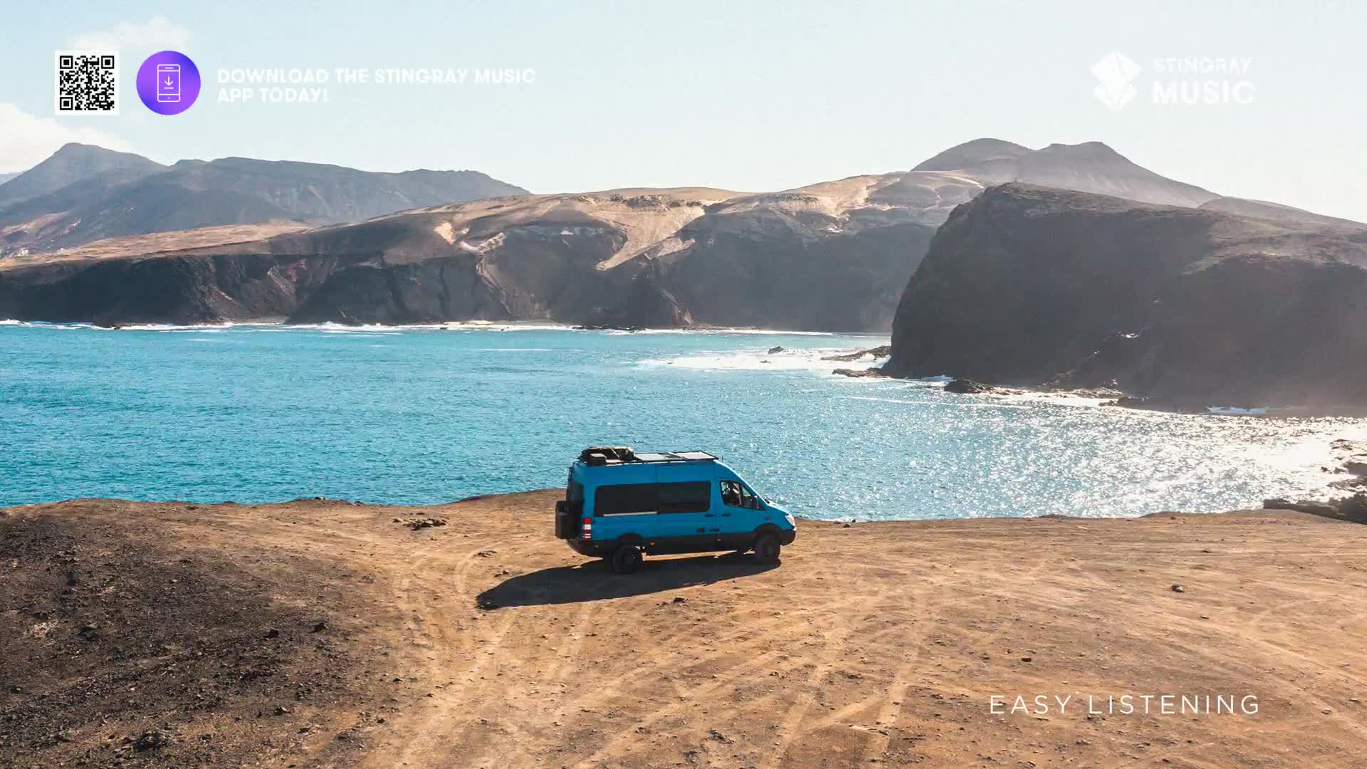 A bright blue camper van sits parked on a dusty, uneven track overlooking a vast expanse of turquoise ocean. Rugged, sun-baked hills rise in the distance, framing the dramatic coastline.