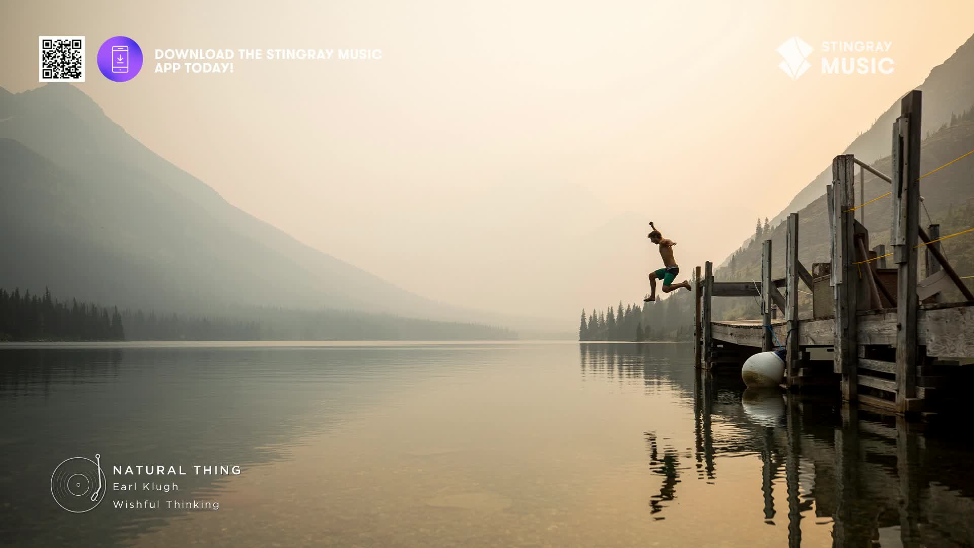 A young person leaps from a wooden dock into a calm, glassy lake. The hazy, mountainous Canadian landscape stretches out behind them, bathed in soft, warm light.