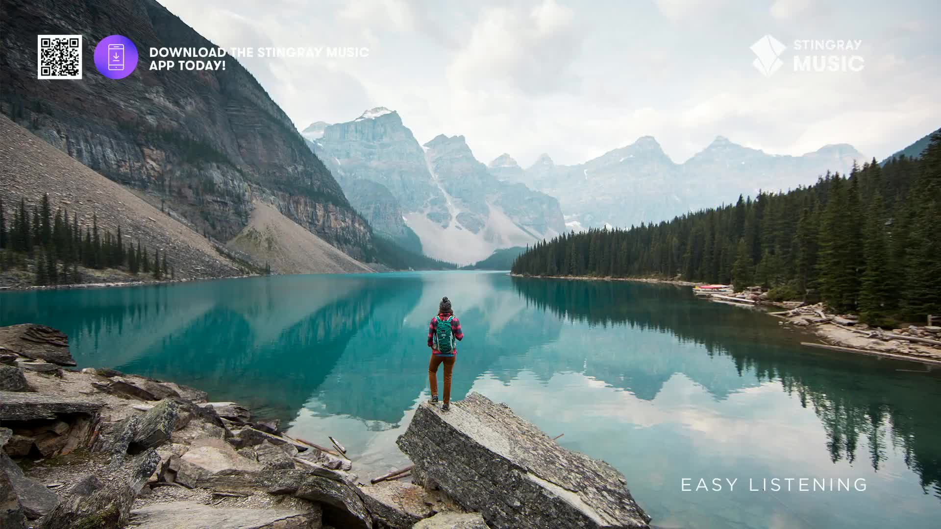 A lone hiker stands on a rock, gazing out at the turquoise waters of Moraine Lake in Banff National Park. The vast Canadian Rockies form a dramatic backdrop, their peaks reflected in the still lake.