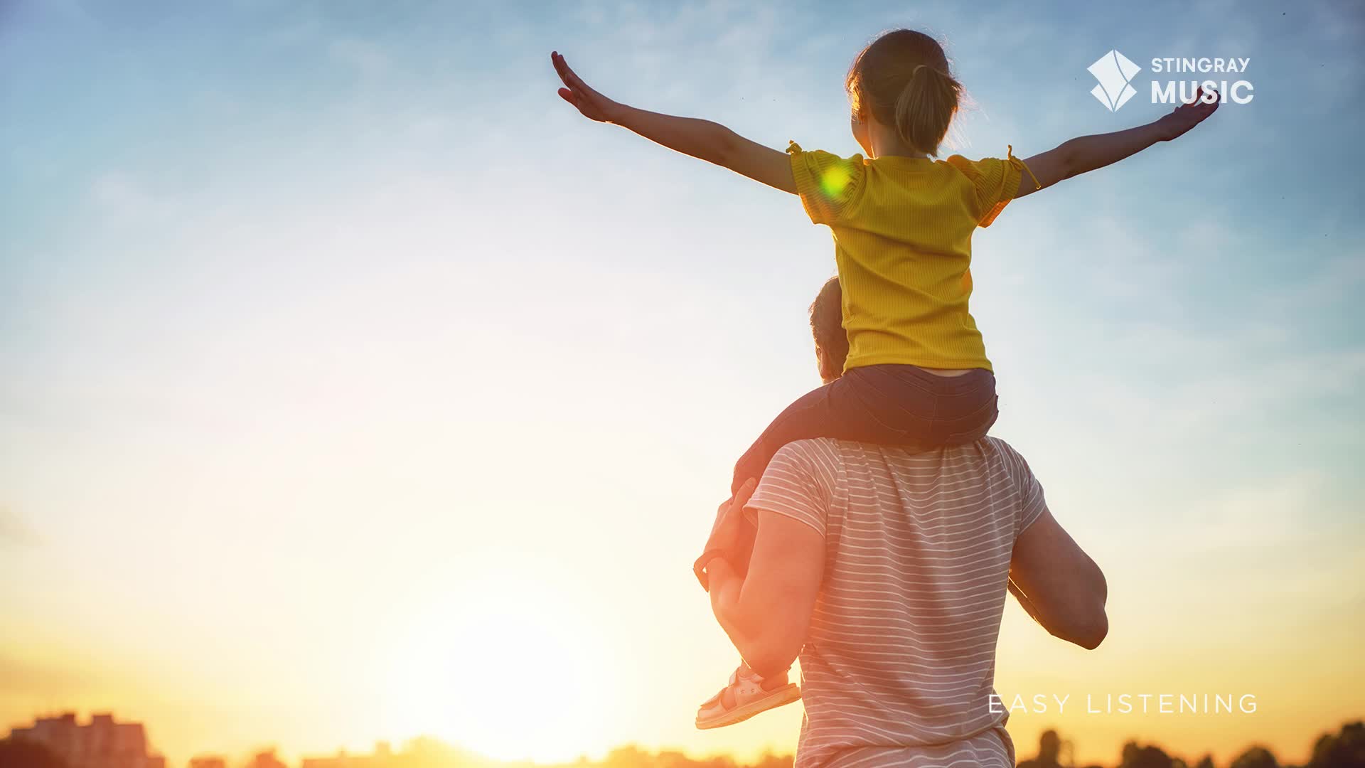 A child with arms outstretched rides on their parent's shoulders, silhouetted against a warm, hazy sunset over a Canadian cityscape. The gentle glow of the setting sun illuminates the scene, creating a peaceful atmosphere.