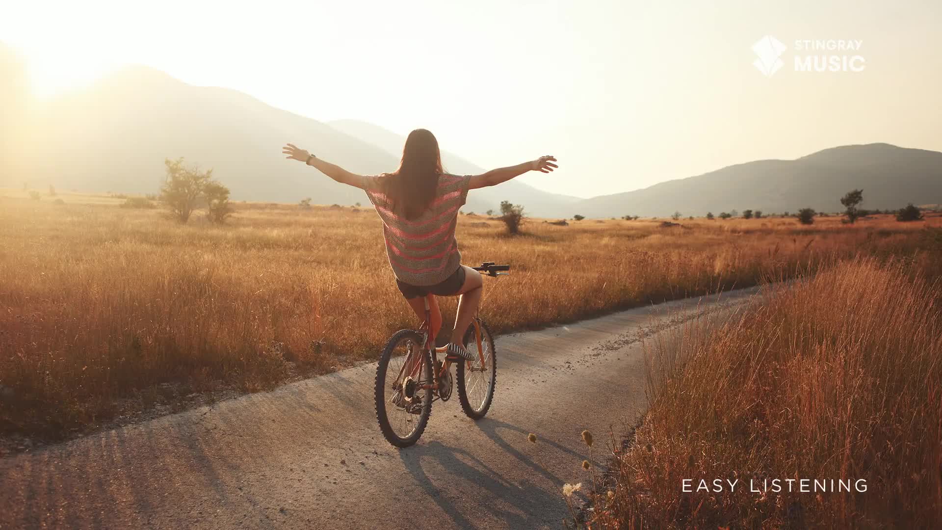 A young woman cycles down a dusty path, her arms spread wide. The late afternoon sun casts a warm glow over the golden fields and distant mountains of this Canadian landscape.
