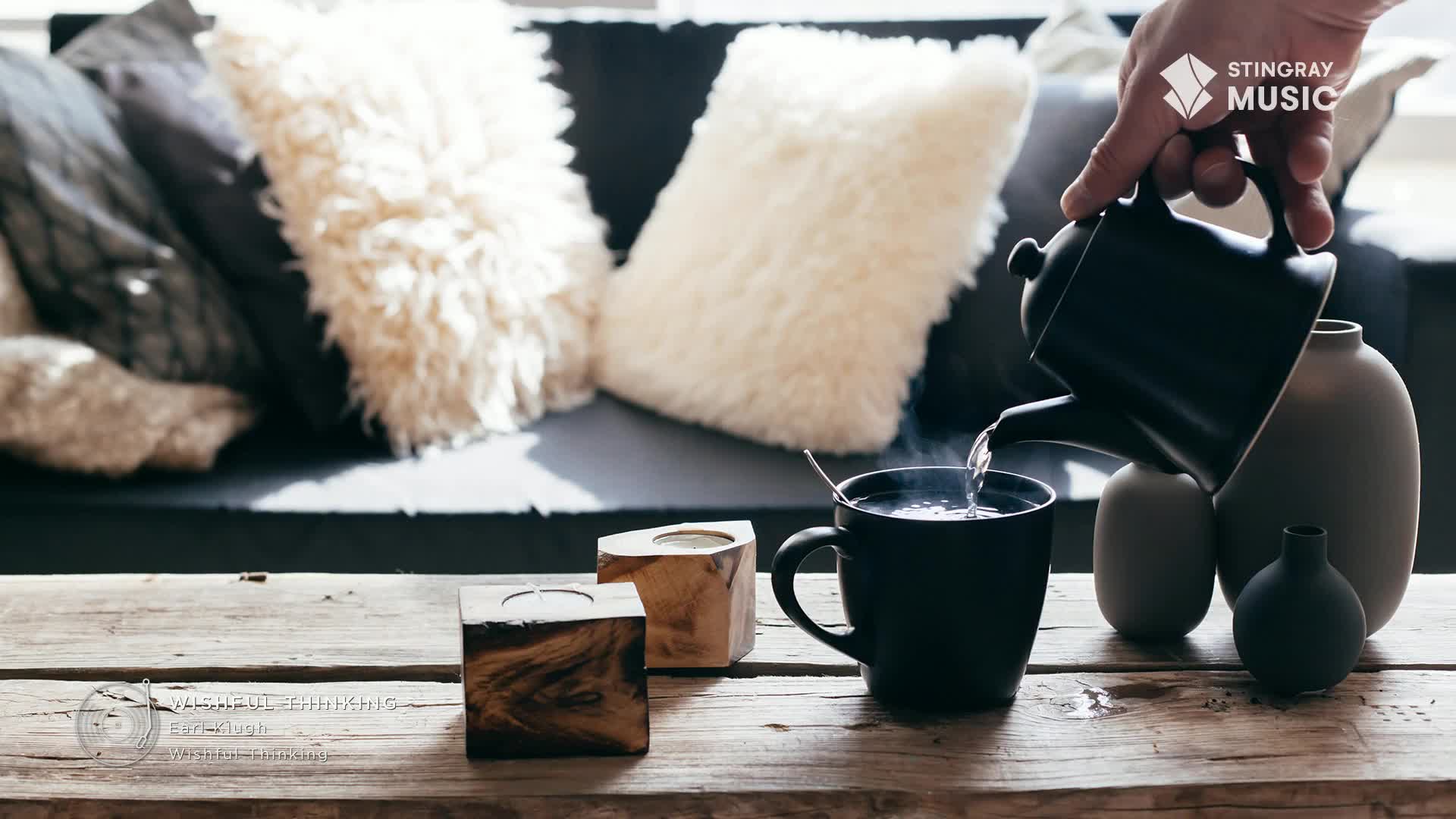 Hot water streams from a black teapot into a dark mug, creating steam that rises into the air. Beside the mug, two wooden candle holders sit on a rustic wooden table.