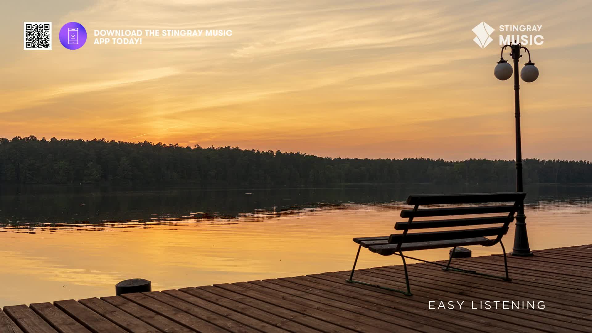 The sun is setting over a calm lake in Canada, casting a warm orange glow across the water. A wooden dock with a bench and lamppost sits empty, waiting for someone to enjoy the peaceful evening.