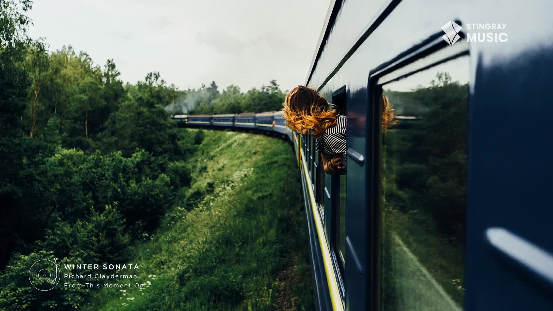 A long blue train snakes through a lush green Canadian landscape. A young woman leans out of a window, her auburn hair catching the light as the train speeds past.