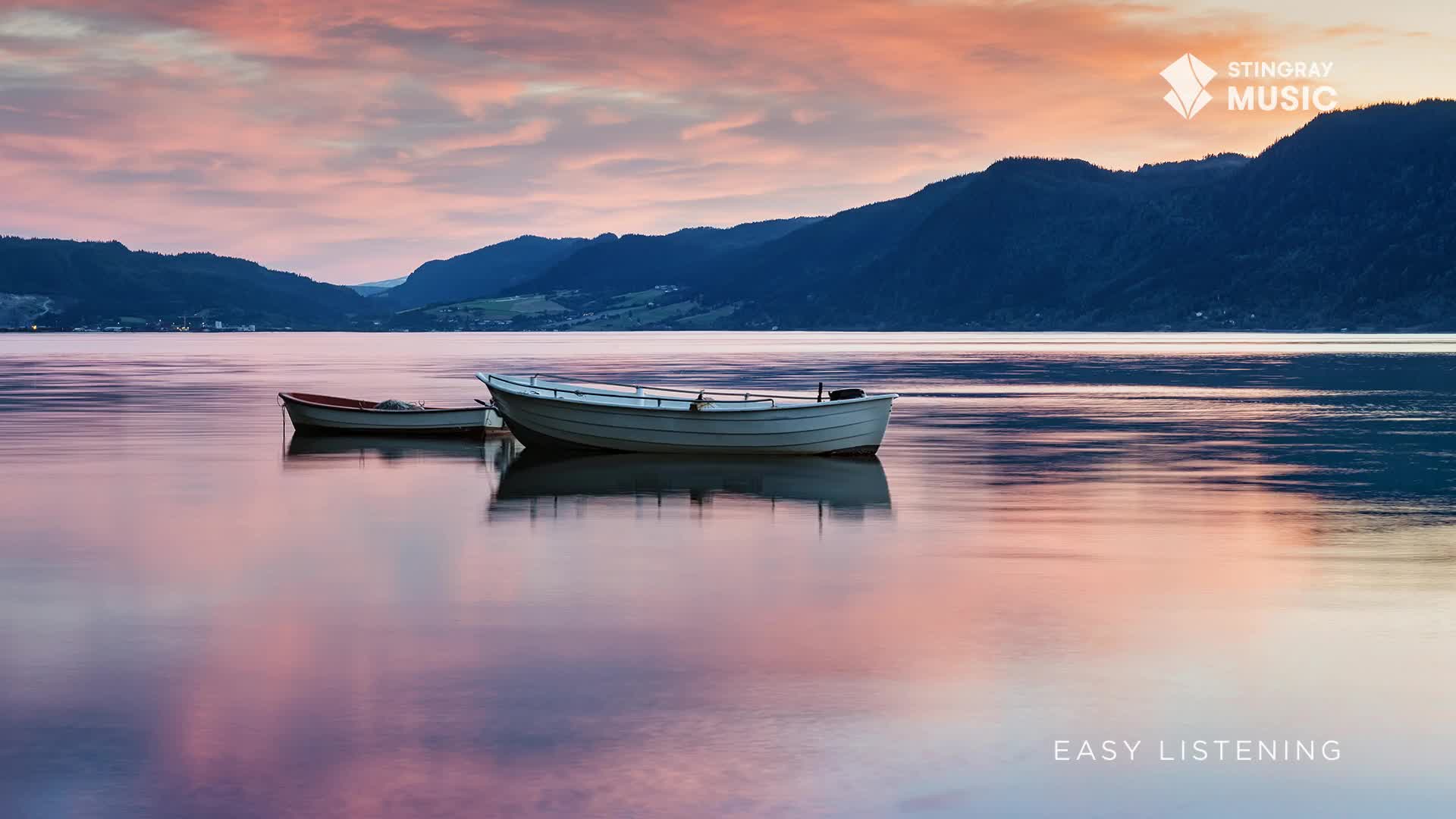 Two small boats rest on the still water, their reflections mirroring the soft pink and orange hues of the sky. The calm lake, framed by dark, distant mountains, feels like a peaceful moment in rural Canada.