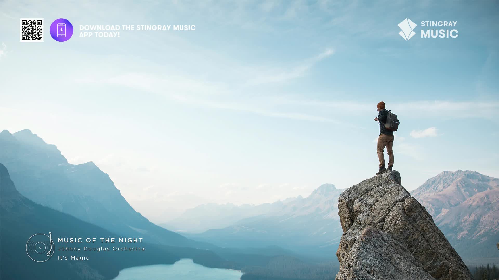 A lone hiker stands on a rocky outcrop, gazing out at a vast Canadian mountain range. The air is clear, and the sky stretches out in a pale blue expanse.