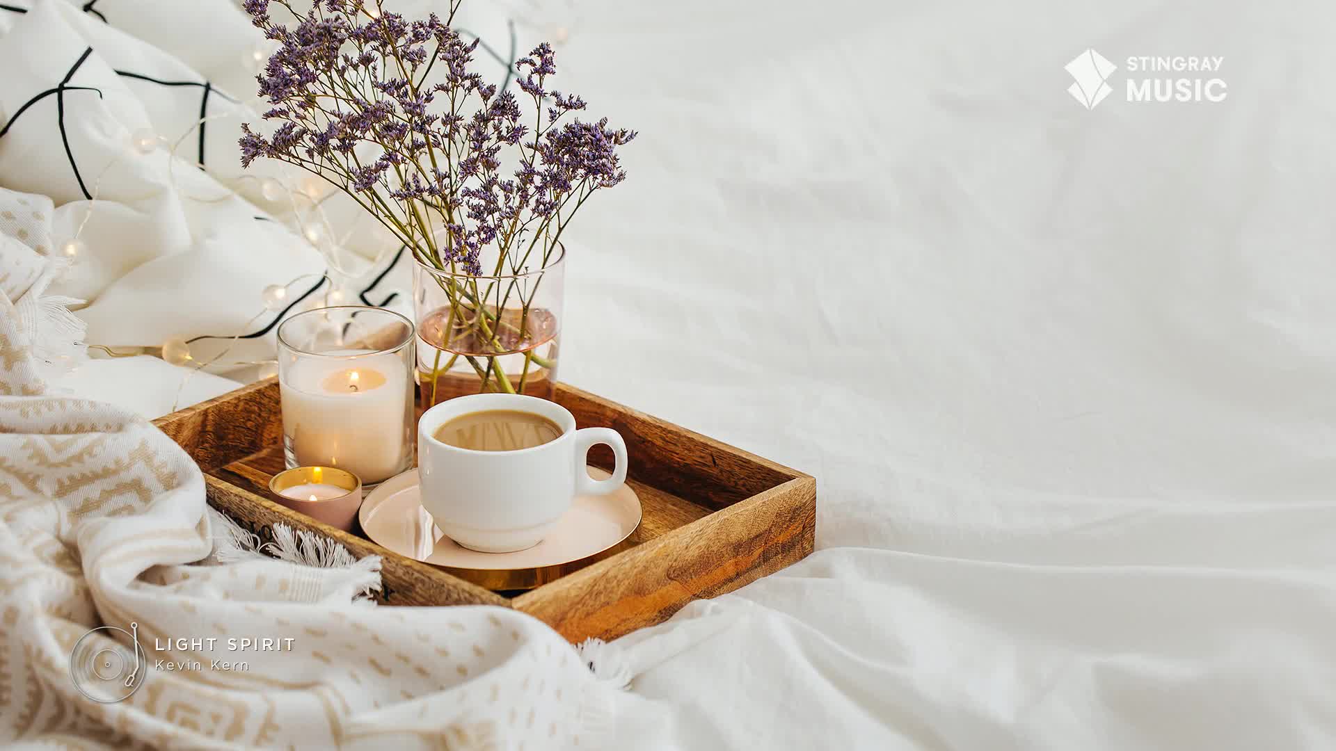 A wooden tray holds a steaming mug of coffee, two lit candles, and a small vase of dried flowers, all nestled amongst soft, white bedding. The gentle glow of the candles and the warm coffee suggest a moment of quiet relaxation, perhaps with some Stingray Easy Listening playing softly in the background.