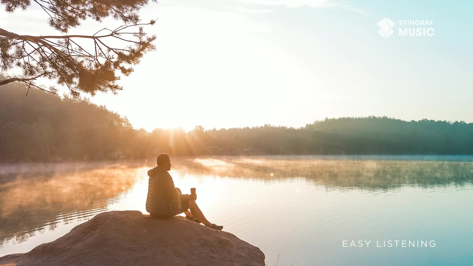 A man sits on a rock, holding a mug, as the sun rises over a misty Canadian lake. The gentle light reflects off the water, creating a peaceful scene.