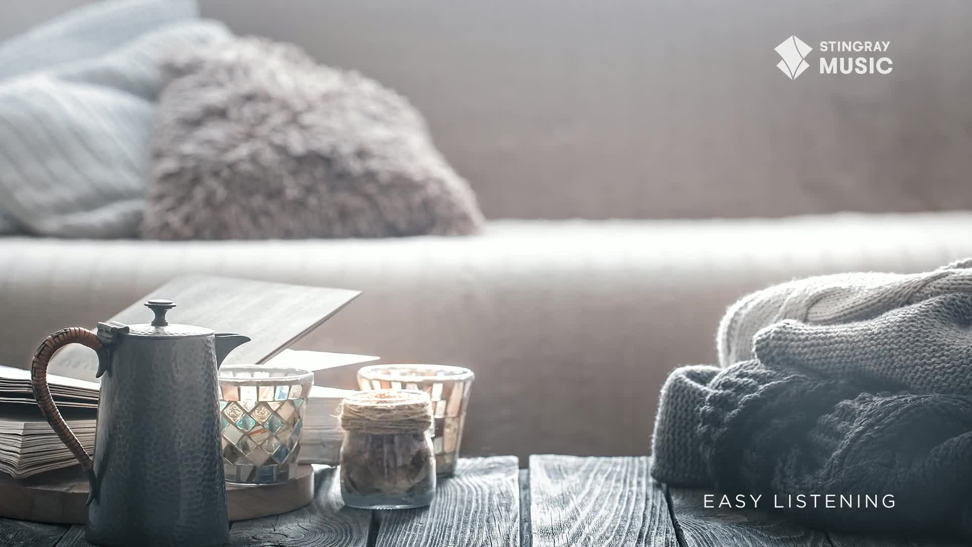 A dark metal coffee pot rests on a wooden table beside a stack of books and decorative candle holders. Nearby, a pile of knitted blankets sits, suggesting a cozy evening in Canada.