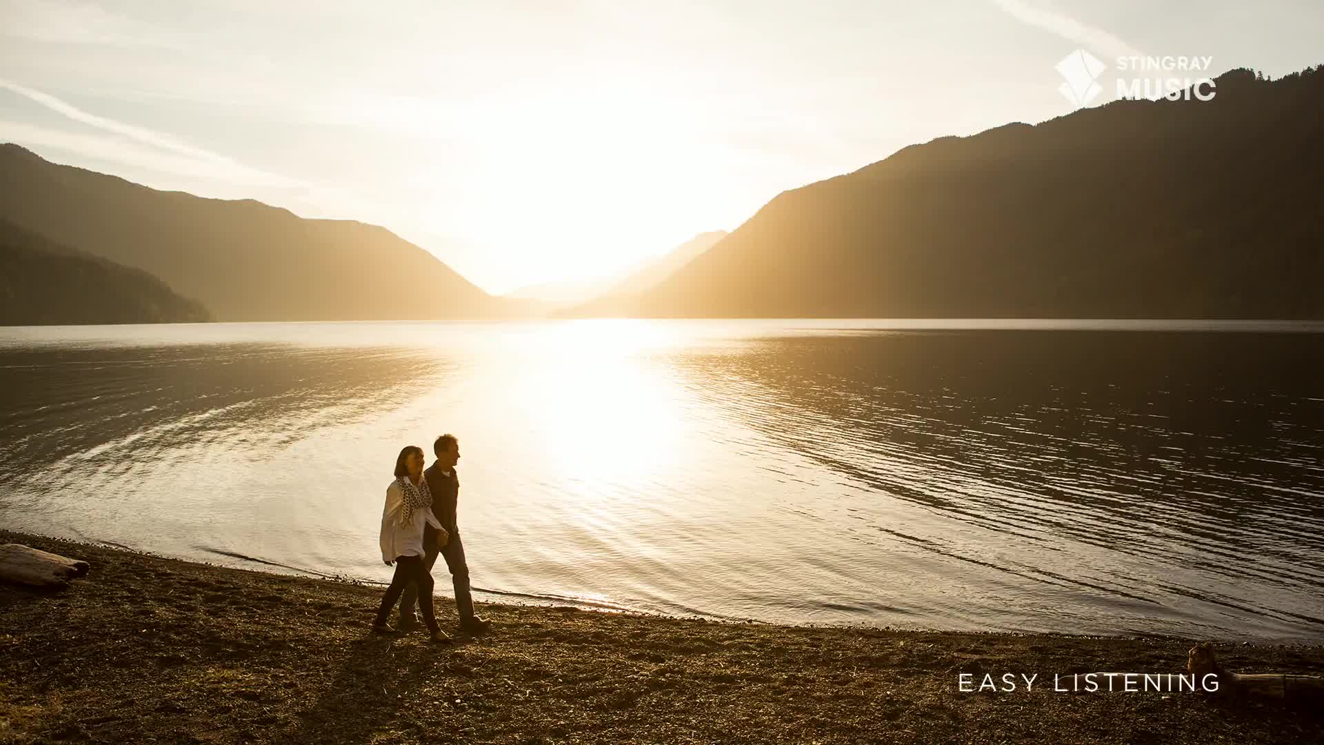 Two people stroll along a pebbly shore as the sun dips behind the mountains, casting a warm, golden light across the calm water. The scene feels like a quiet moment in the Canadian wilderness, a perfect soundtrack for Stingray Easy Listening.