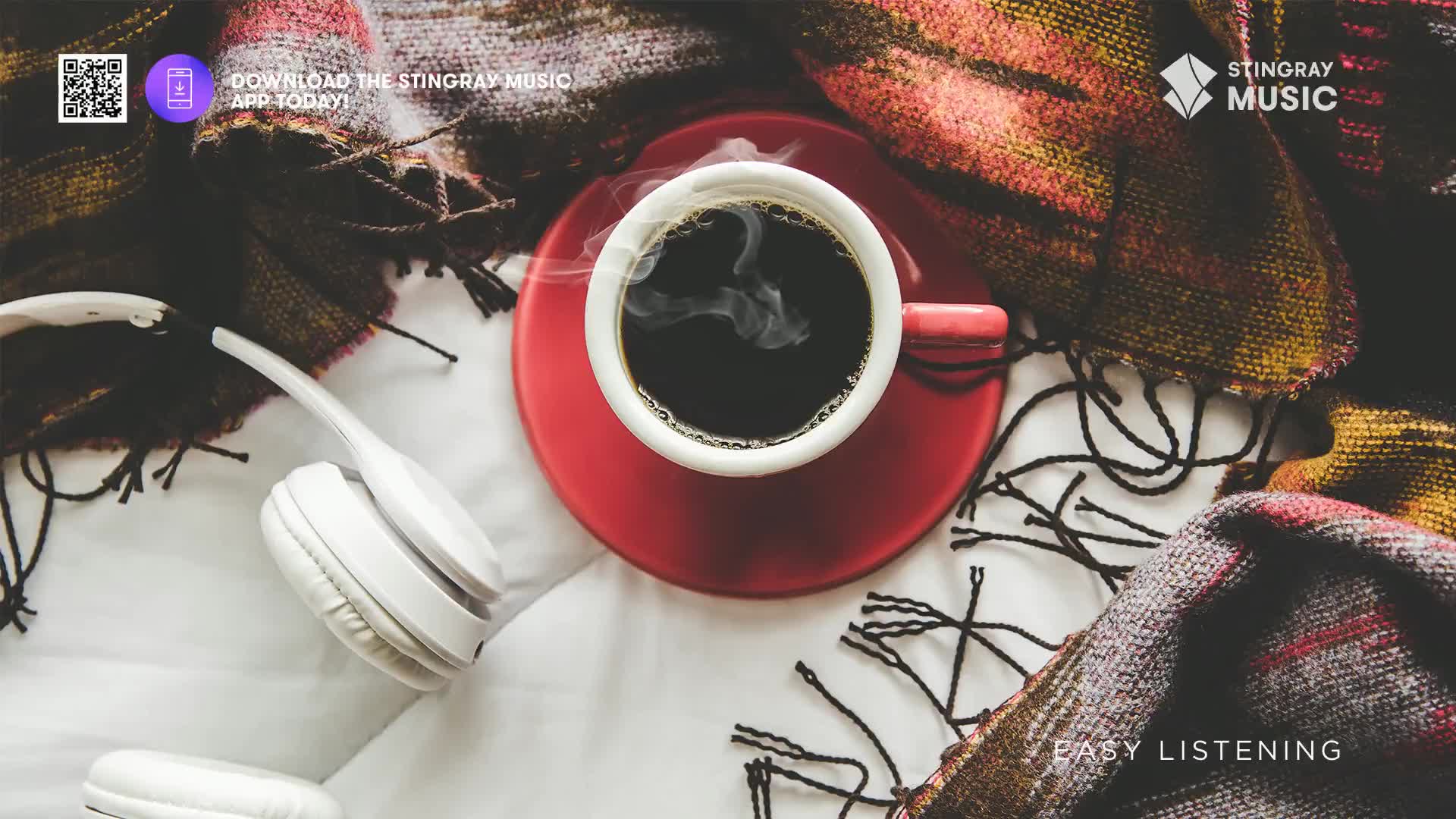 Steam rises from a fresh cup of coffee, nestled beside a pair of white headphones. A cozy, patterned blanket is draped around the scene, suggesting a quiet moment.