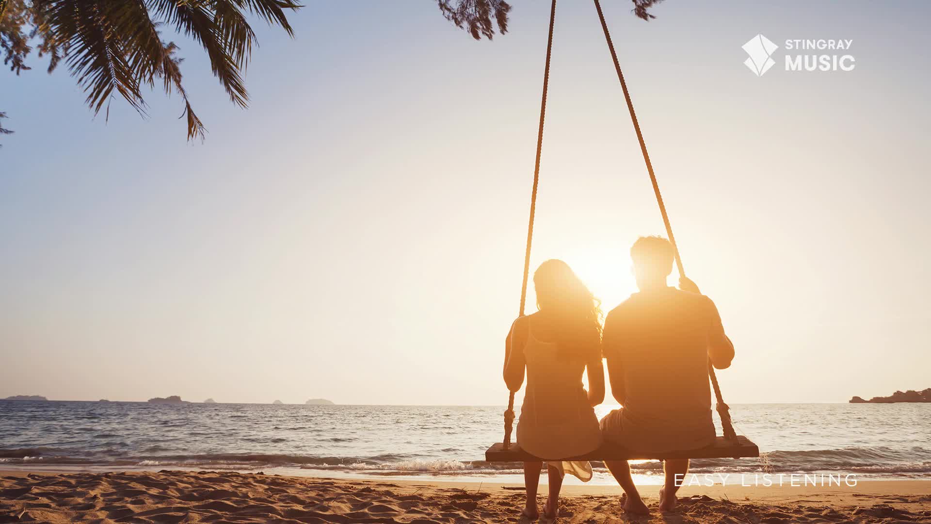 A couple sits on a swing, their backs to the ocean. The sun dips low, casting a warm glow over the water.