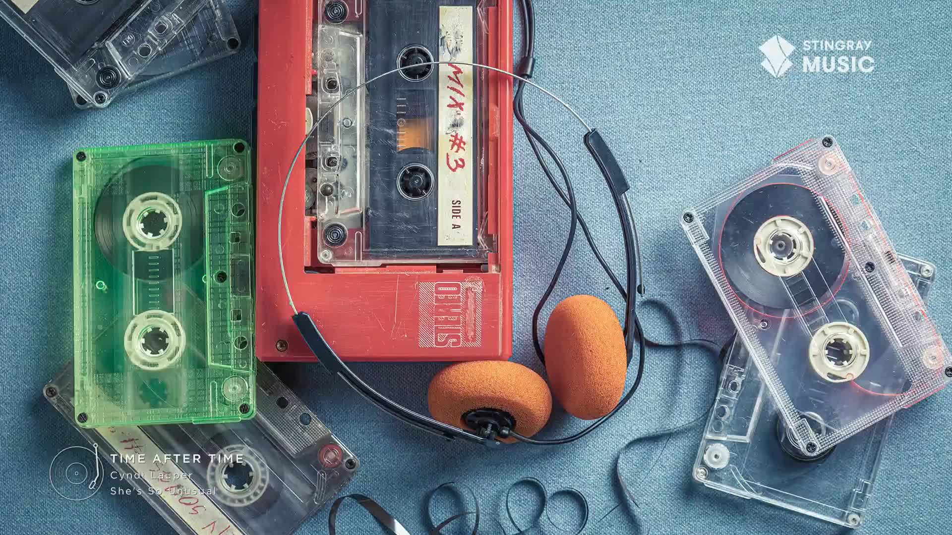 A red cassette player sits open, a "Mix #3" tape inside, and headphones draped over the top. Scattered around are several clear cassette tapes, some green-tinted, all on a blue fabric, likely from a Stingray Everything '80s playlist.
