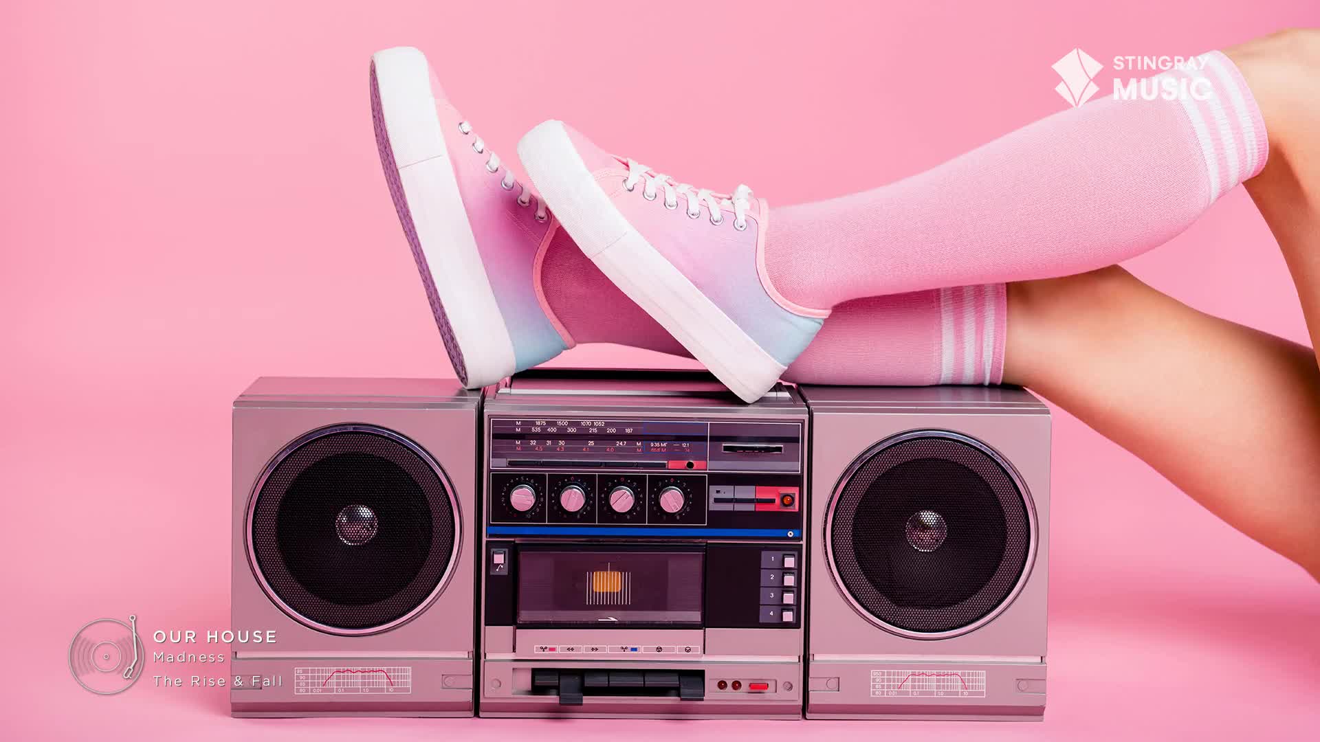 A boombox sits on a pink background with a pair of legs resting on top. The legs are clad in pink socks and sneakers, the latter with a gradient of pink, purple, and blue.
