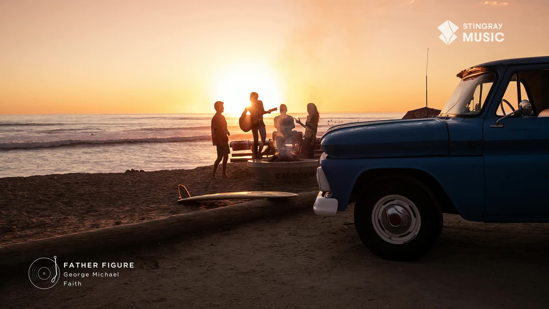 The sun dips low, casting long shadows as a group gathers around a beach bonfire. A vintage blue truck sits parked nearby, a surfboard resting on the sand.
