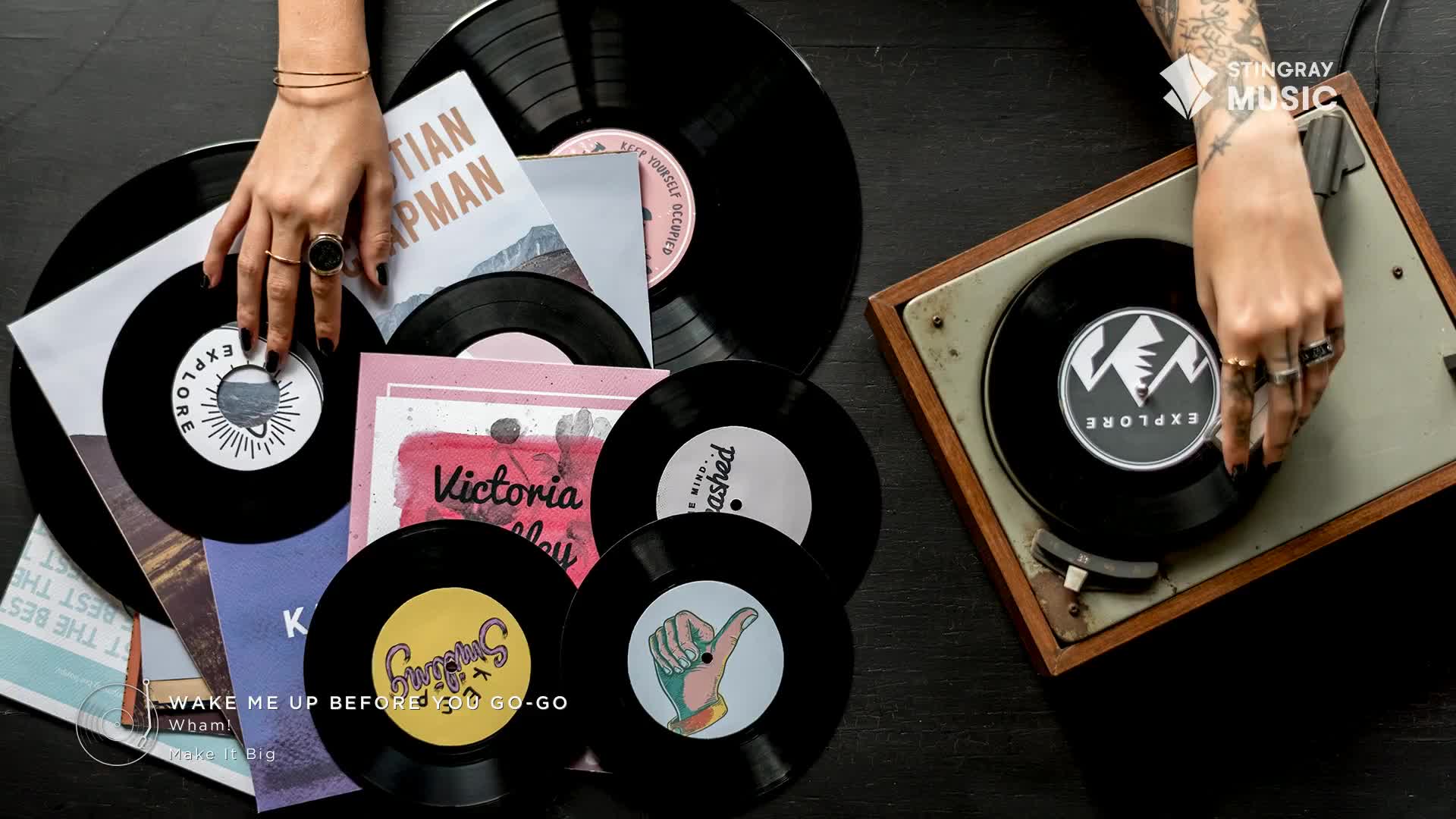 A hand gently lowers a black vinyl record onto an old turntable. Other records, with colourful labels and album art, are scattered around.
