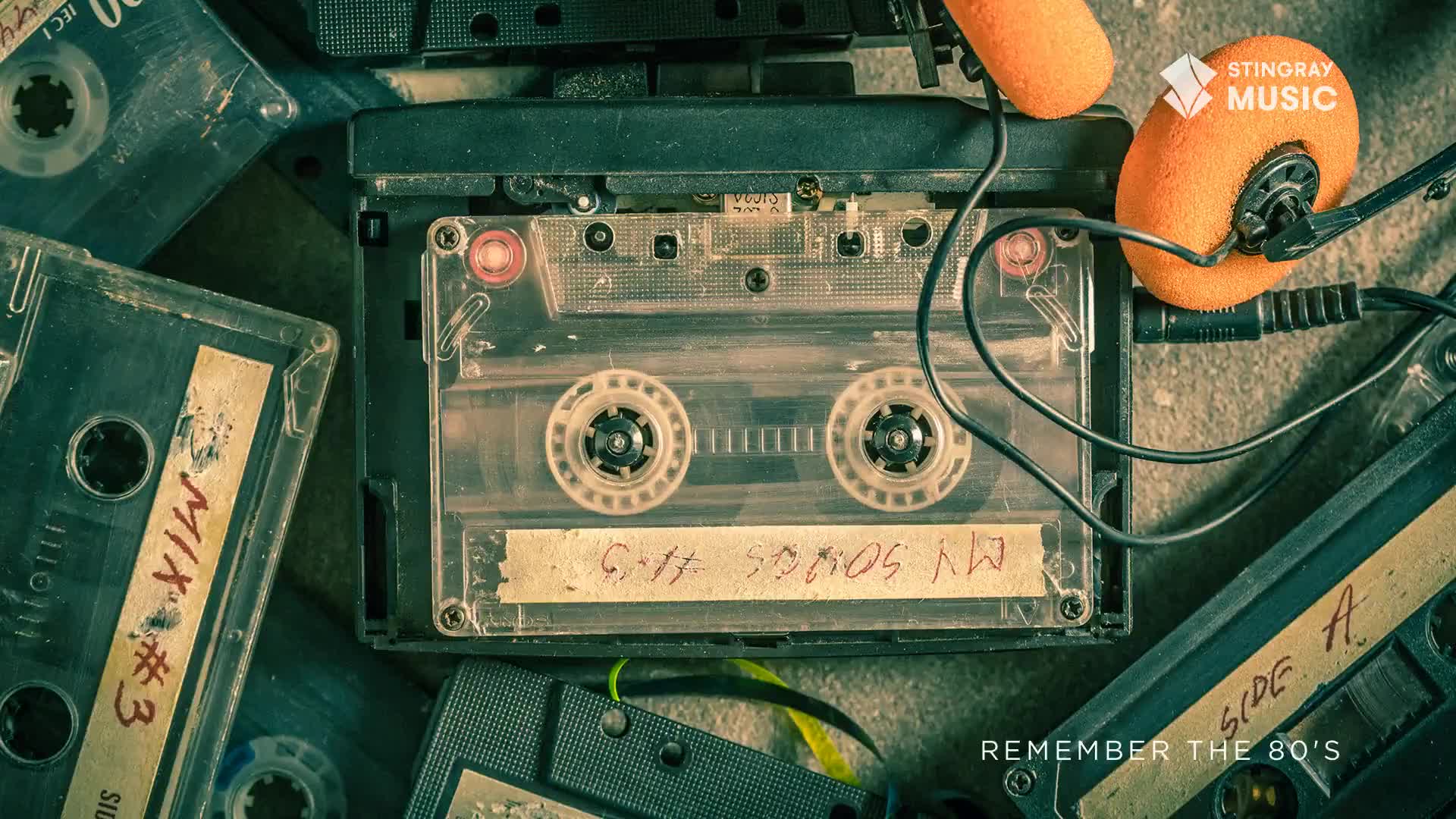A cassette player sits surrounded by other tapes, with orange headphones plugged in. The clear tape in the player has a handwritten label, a reminder of Canada's Stingray Everything '80s.
