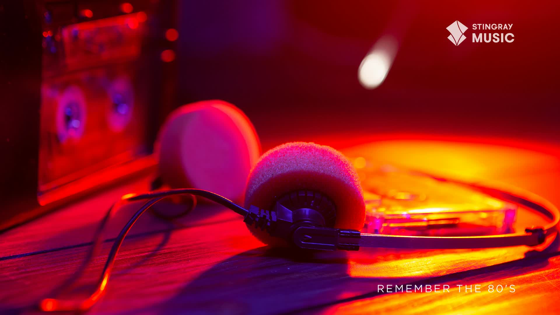 Headphones with foam earcups rest on a wooden surface bathed in red and orange light. A cassette player and a compact disc player are visible in the background, evoking Stingray Everything '80s from Canada.
