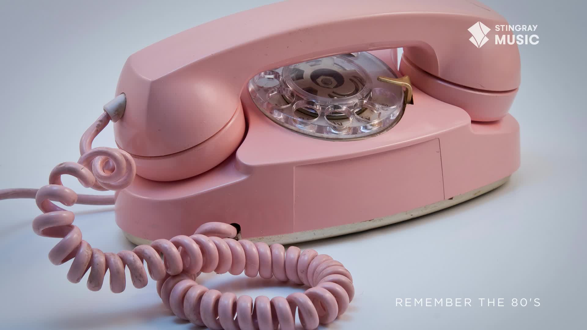 A vintage pink rotary phone sits on a white surface, its coiled cord snaking across the frame. The Stingray Music logo and the text "Remember the 80's" hint at a nostalgic vibe, probably from Canada.
