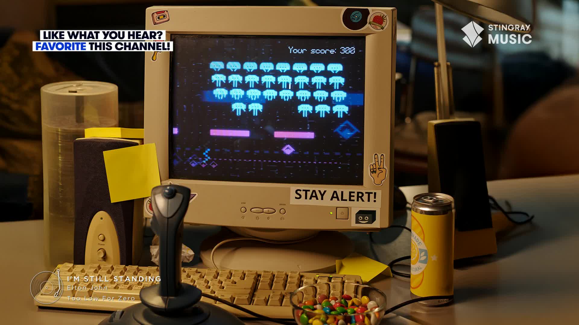 A retro computer screen displays an alien invasion game, with a joystick ready for action.  A Stingray Music logo and the words "I'm Still Standing" suggest a nostalgic vibe, maybe the '80s.
