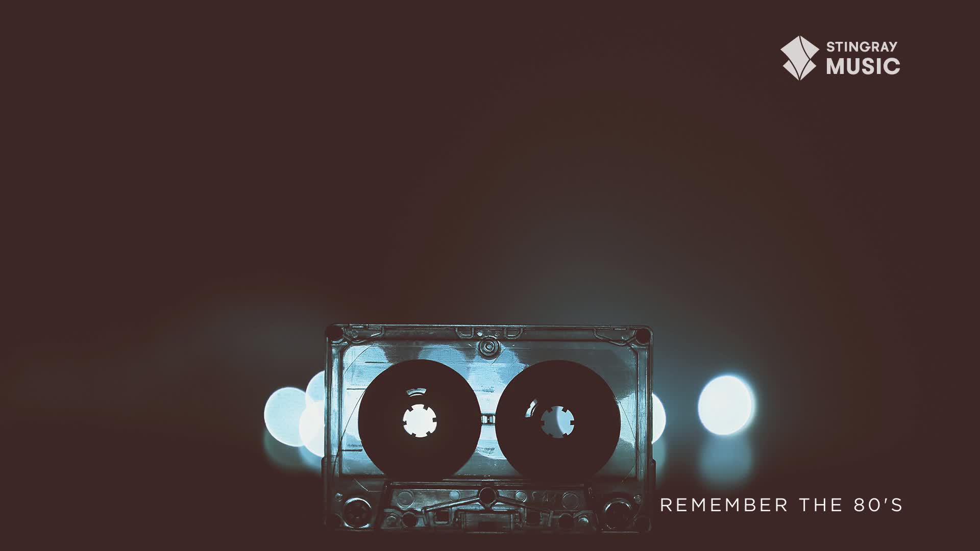 A clear cassette tape sits centered, its dark reels contrasting against the soft, out-of-focus lights. The Stingray Music logo and "Remember the 80's" text are visible, a nostalgic nod to the music of Canada's past.
