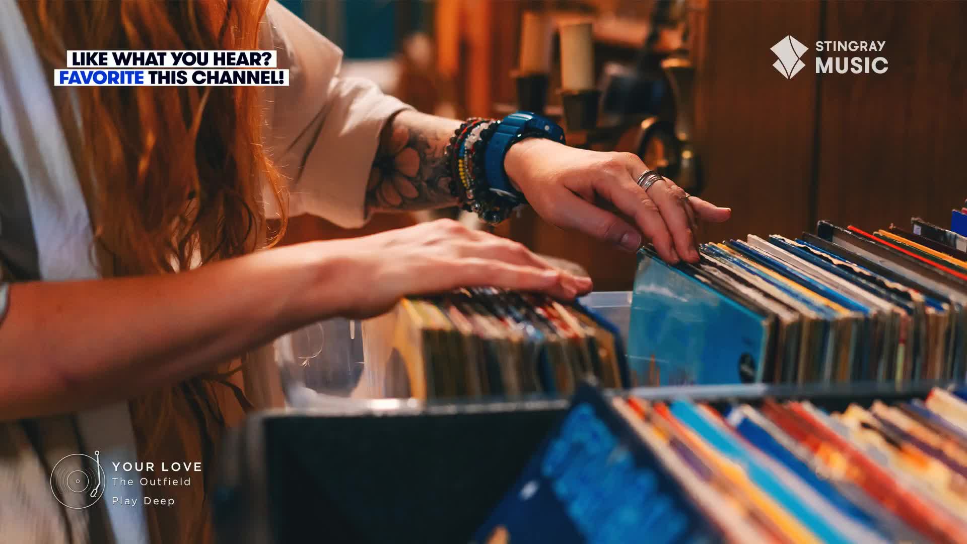 A hand with multiple bracelets and rings carefully moves through a stack of vinyl records. The person is likely in Canada, enjoying Stingray Everything '80s, as "Your Love" by The Outfield plays.
