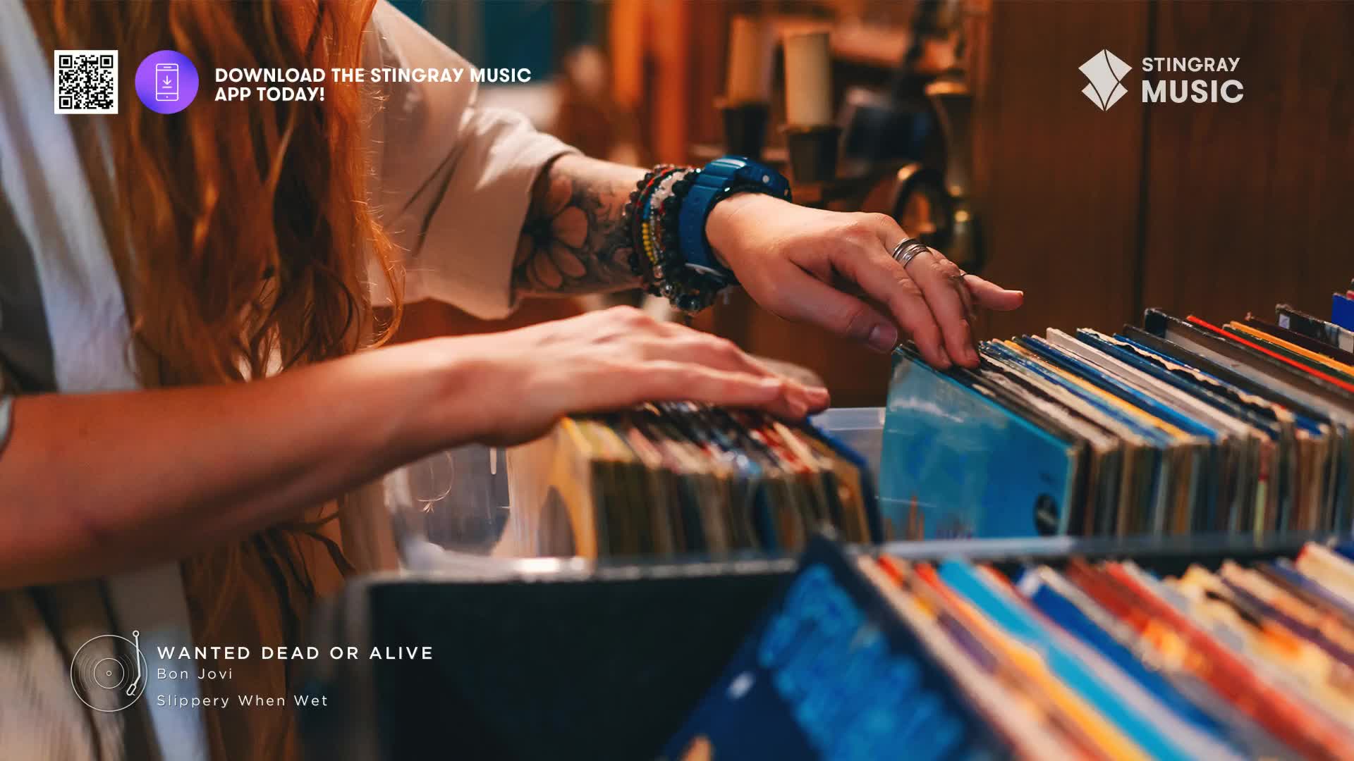 A hand with multiple bracelets and a tattoo gently flips through a stack of vinyl records. The album covers are a mix of colors, and the scene feels like a visit to a record store, perhaps in Canada.
