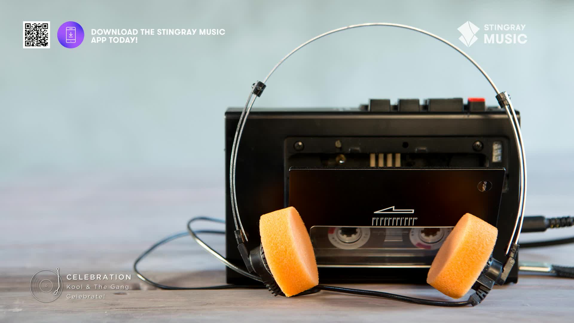 A black cassette player with orange foam headphones sits on a wooden surface. The player's cassette door is open, revealing the tape inside.
