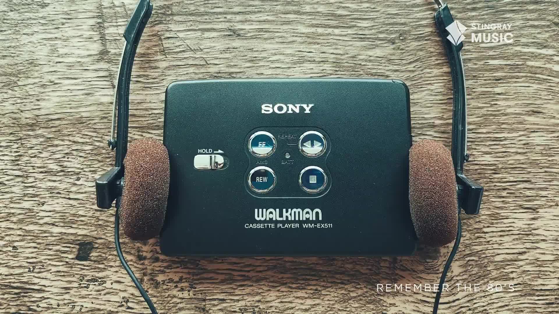 A black Sony Walkman sits on a wooden surface, its buttons and the "HOLD" switch clearly visible. The headphones, with their brown foam pads, are draped on either side.
