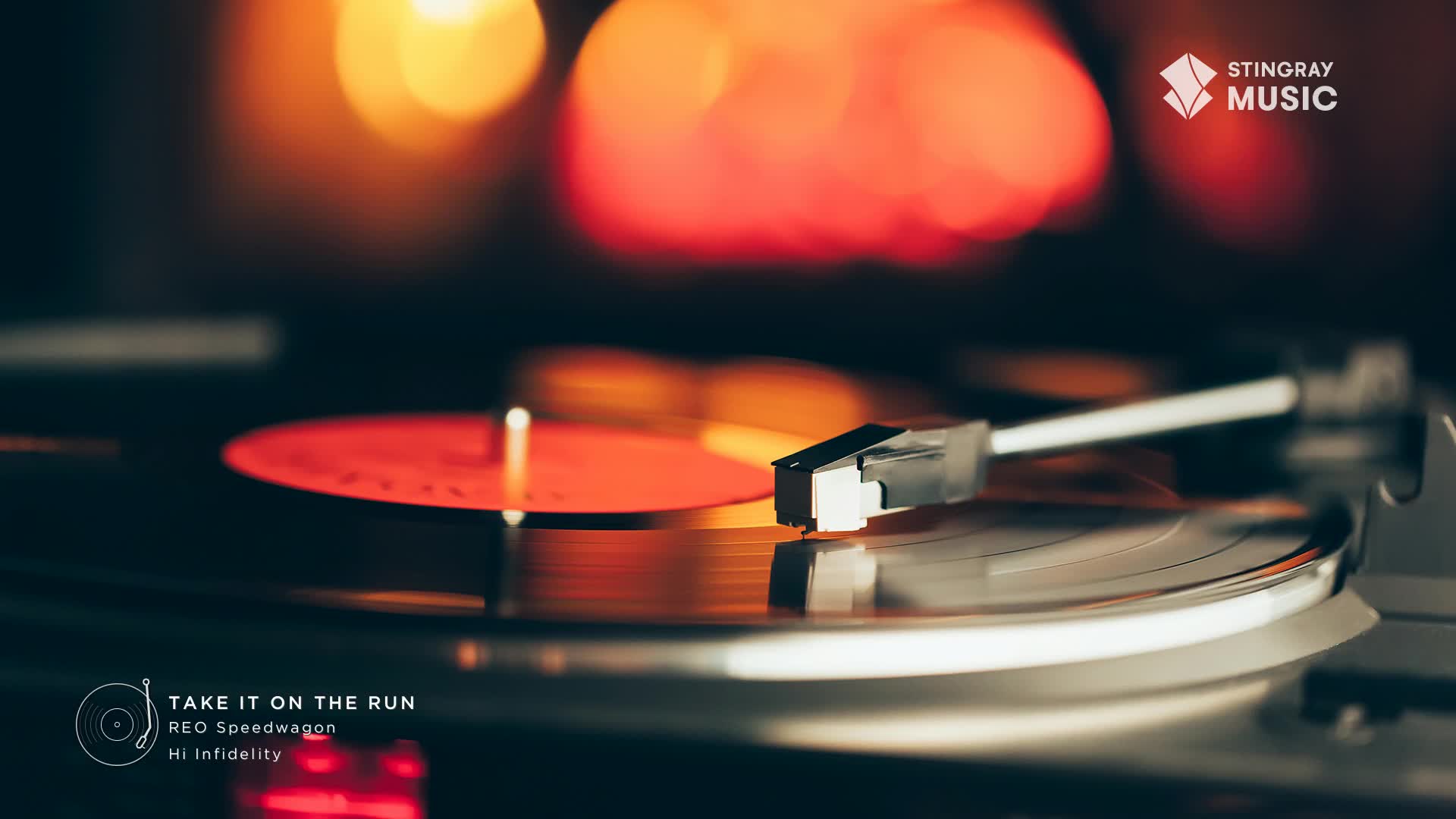 The needle rests on a red vinyl, ready to play. The record spins, reflecting the warm glow of the background lights.
