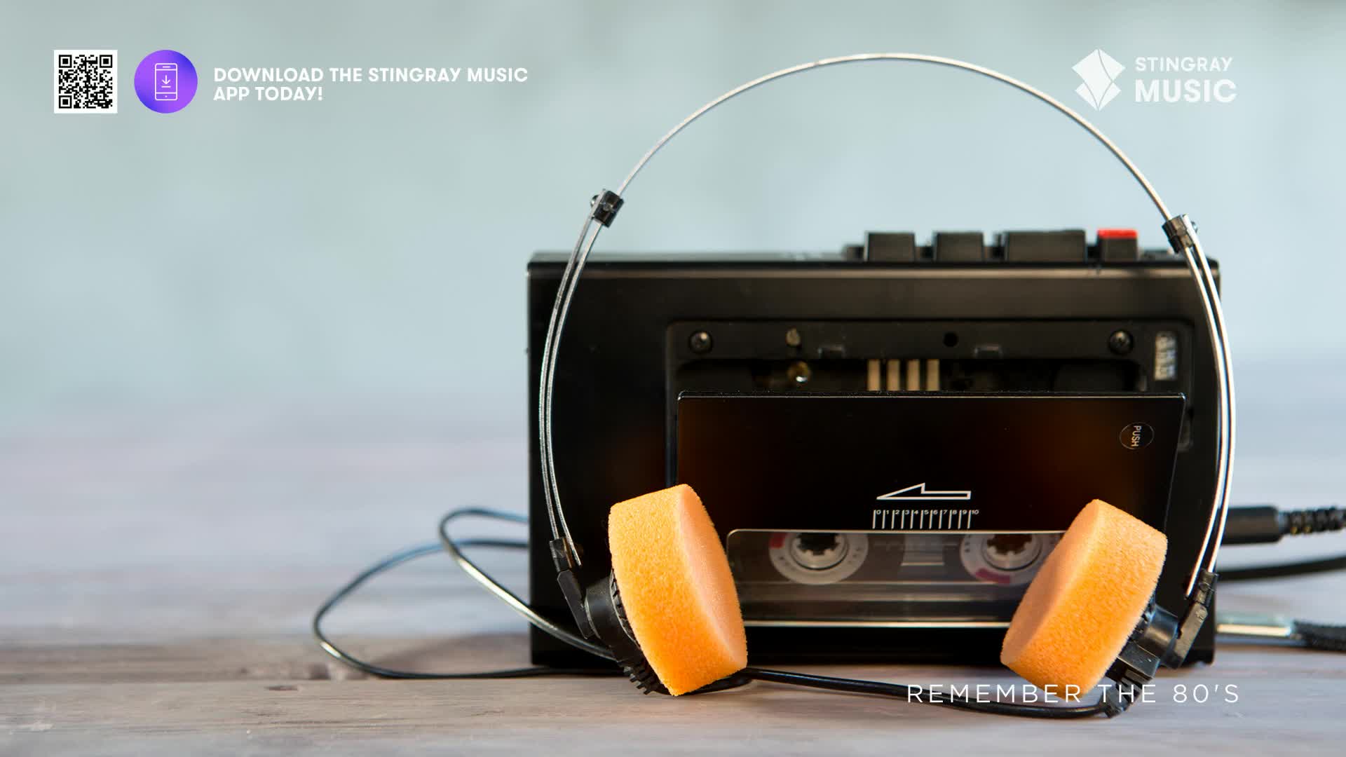 A black cassette player sits on a wooden surface, its headphone jack plugged in. The bright orange ear pads of the headphones rest against the cassette player, ready for some Stingray Everything '80s.
