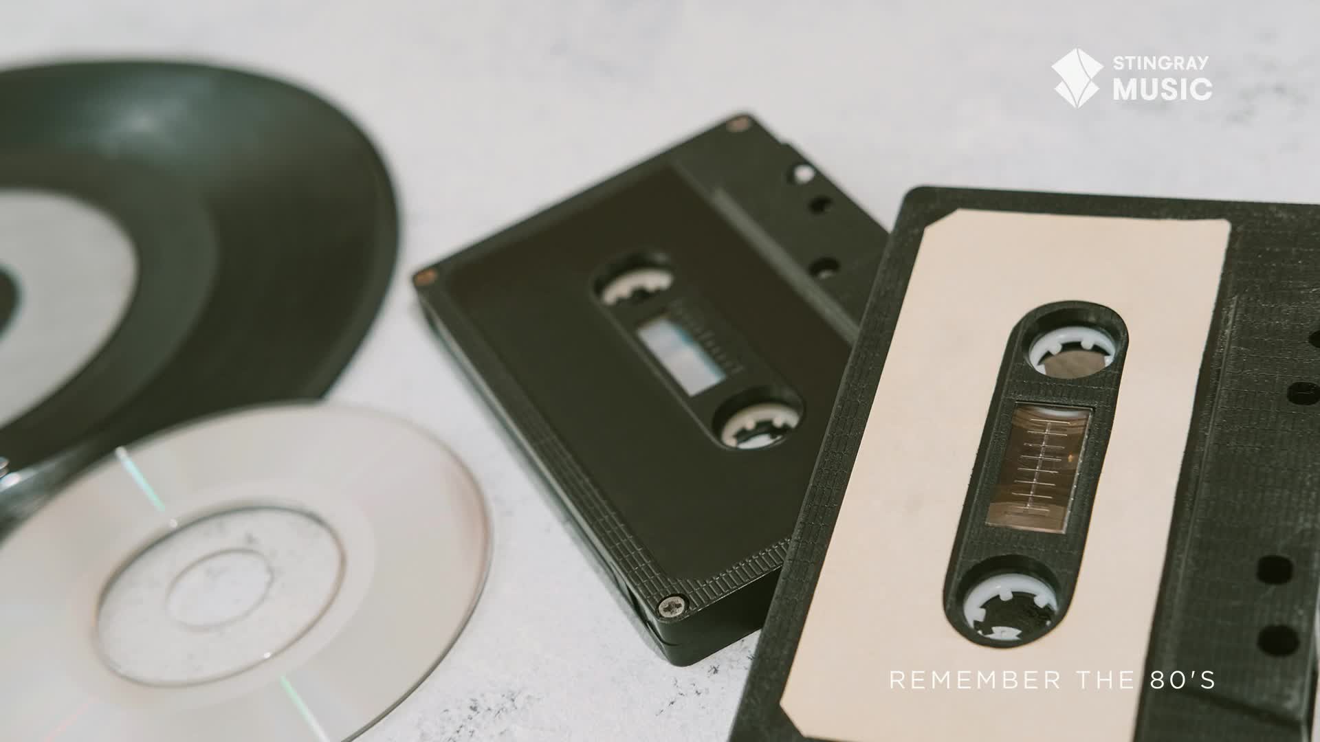 A black cassette tape and a CD sit next to a record, all on a white surface. The Stingray Music logo and "Remember the 80's" text appear in the corner.
