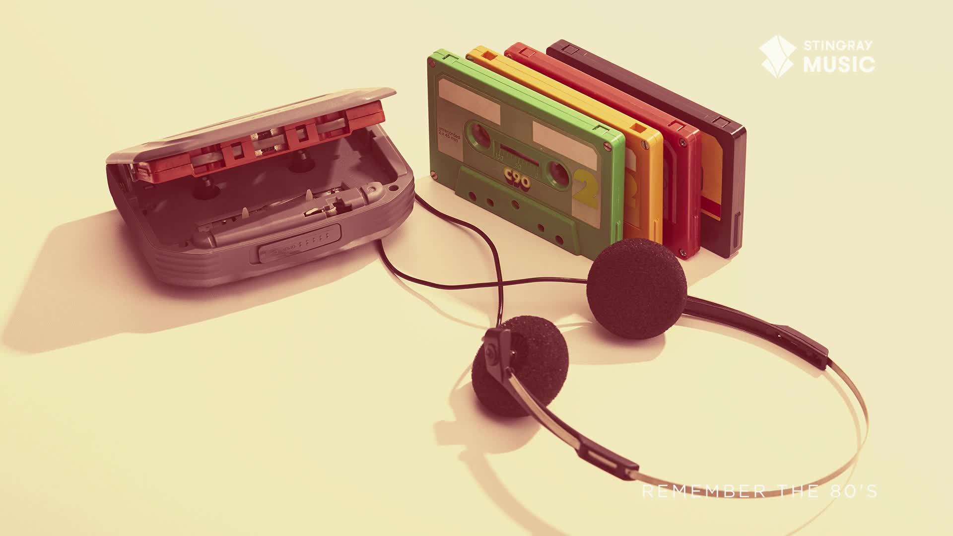 A portable cassette player with its lid open sits beside a row of brightly colored tapes. A pair of black foam-covered headphones rests in front of them, ready for listening.