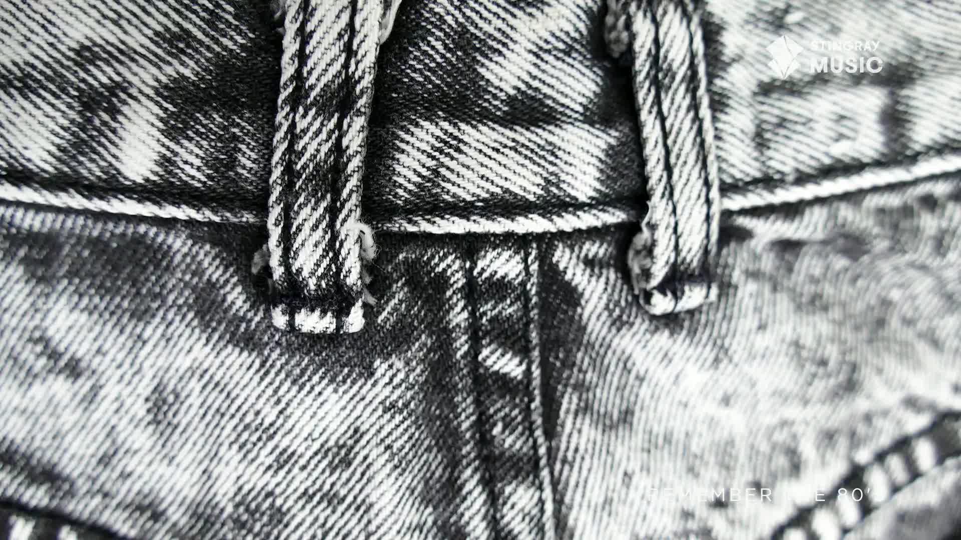 The acid-washed denim of a pair of jeans is in focus, the thick white stitching of the belt loops and fly stand out. A faint "Stingray Everything '80s" logo is visible in the lower right corner.