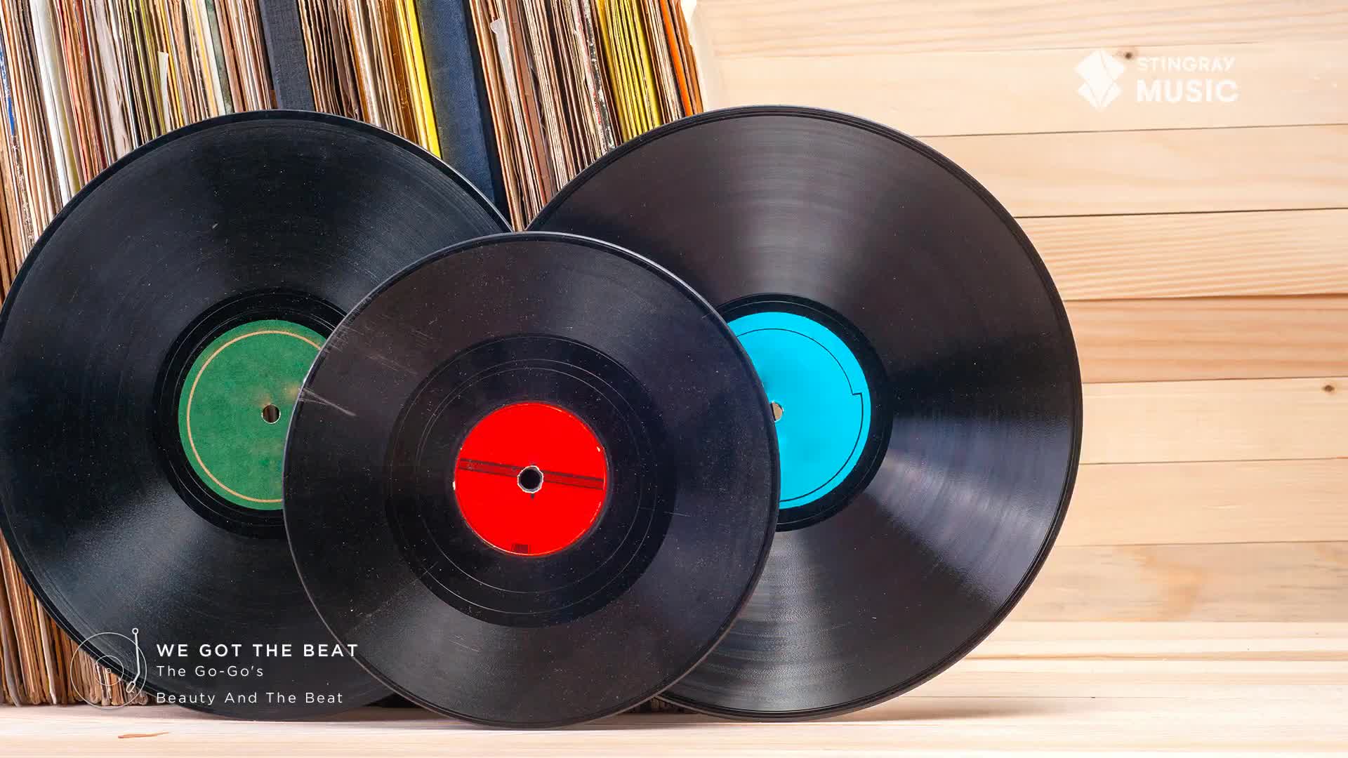 A stack of records leans against a wooden background, with three black vinyl discs in the foreground. One record has a green label, another a red, and the third a bright blue. A stack of records leans against a wooden background, with three black vinyl discs in the foreground. One record has a green label, another a red, and the third a bright blue.