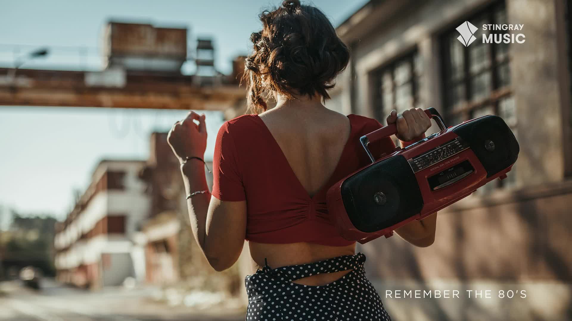 A woman in a red top and polka dot skirt walks away, carrying a red boombox. The industrial buildings behind her suggest a Canadian setting, and the boombox evokes the spirit of the 80s.
