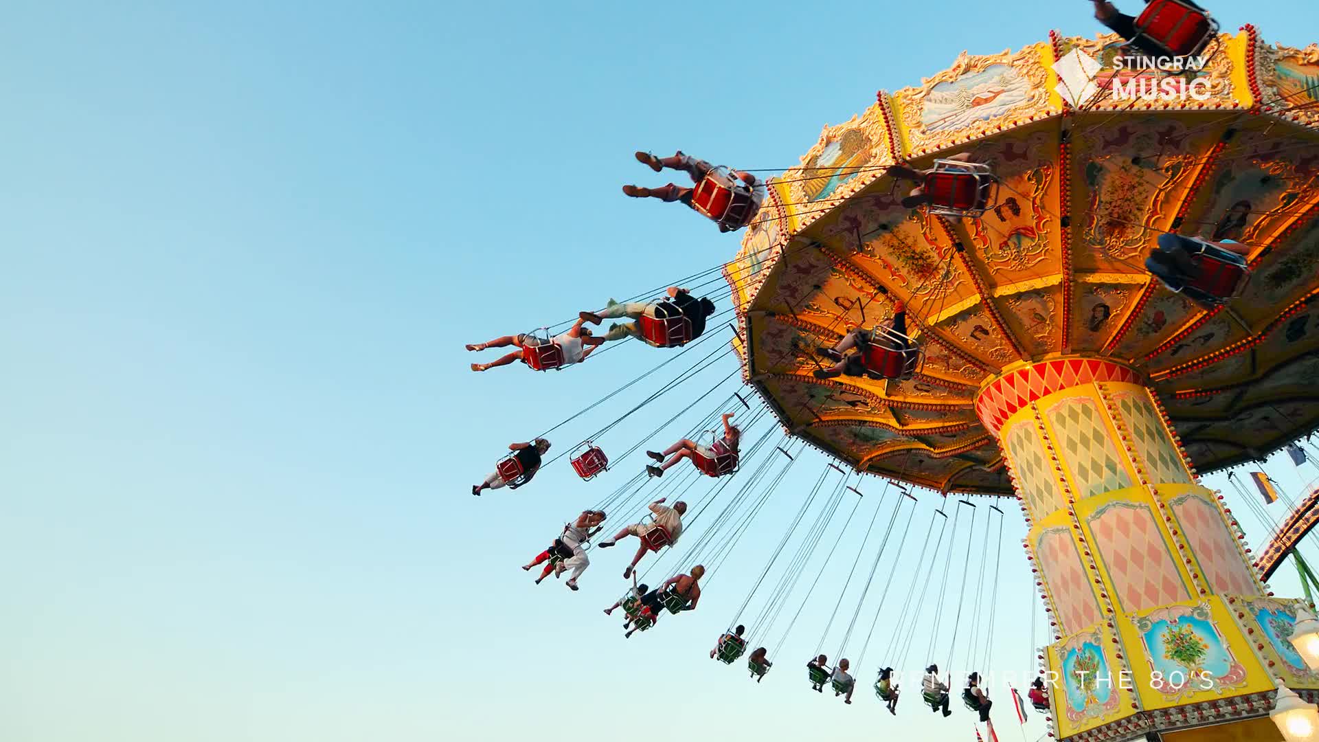 The swings are lifting people high into the bright Canadian sky, their legs dangling as they arc outward. The carousel's ornate, sun-drenched canopy spins above, a vibrant echo of Everything '80s.