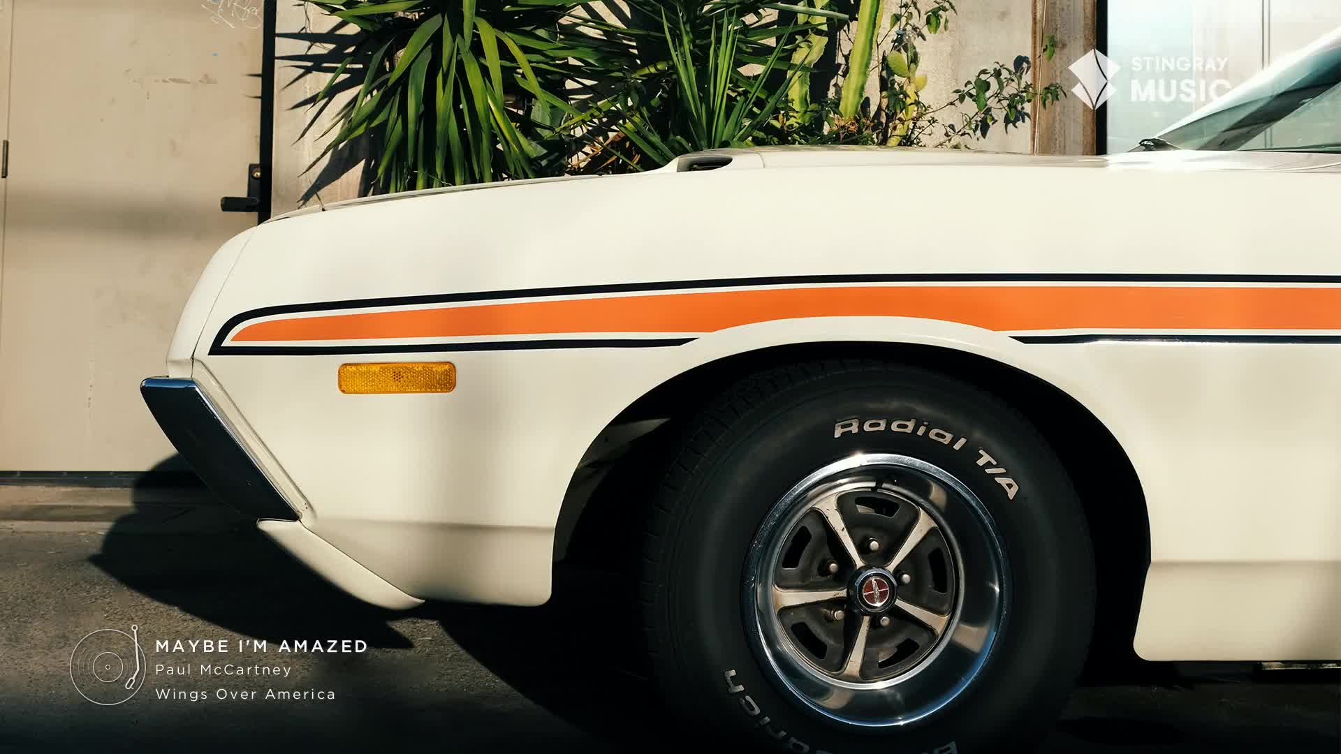 The front quarter of a classic white car, with an orange stripe, sits in the sunlight. A black tire with white lettering and a chrome hubcap is parked, ready for a drive, in what looks like Canada, perhaps during the Stingray Flashback 70's.

