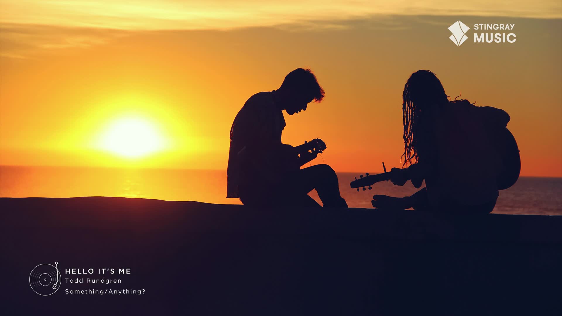 Two figures, silhouetted against the sunset, sit side-by-side, each strumming a guitar. The sky is a warm orange, and the ocean stretches out behind them.
