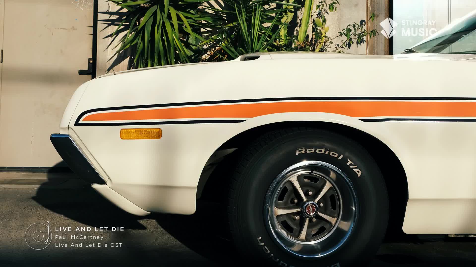 A vintage white car with an orange stripe sits parked, its tire gleaming in the sunlight. The Stingray Flashback 70's vibe is strong, and the "Live and Let Die" soundtrack feels right.
