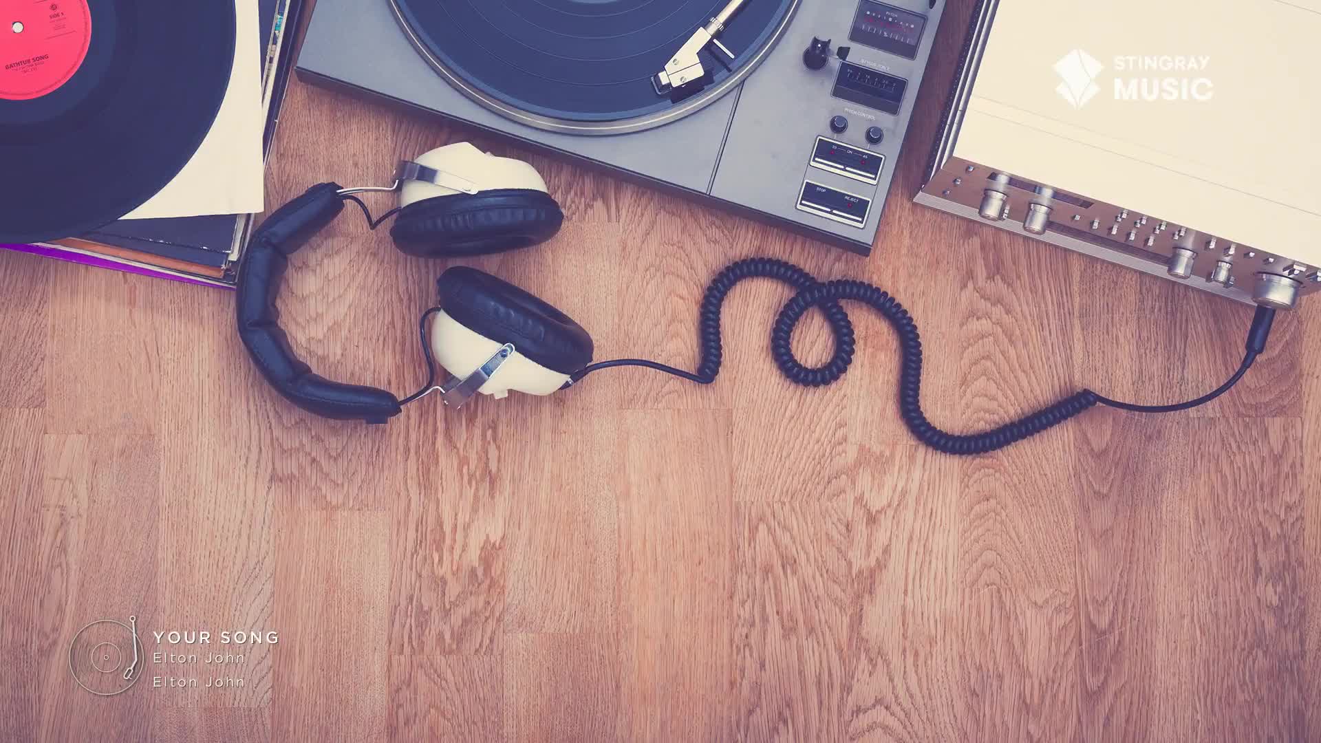 A turntable sits ready, with a vinyl record stacked next to it. White headphones with black earcups rest on the wooden floor, their coiled cord leading to the receiver.
