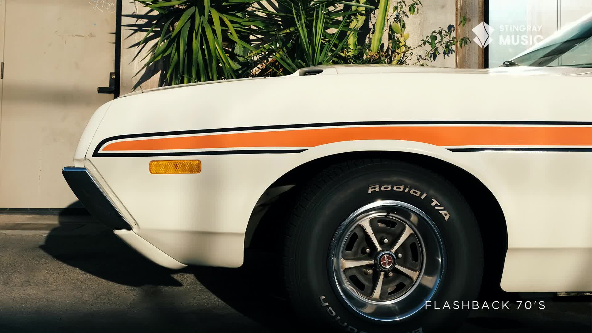 The front quarter of a white car with an orange stripe sits parked, its black tire and chrome wheel reflecting the sunlight. It's a Stingray Flashback from the 70s, probably somewhere in Canada.
